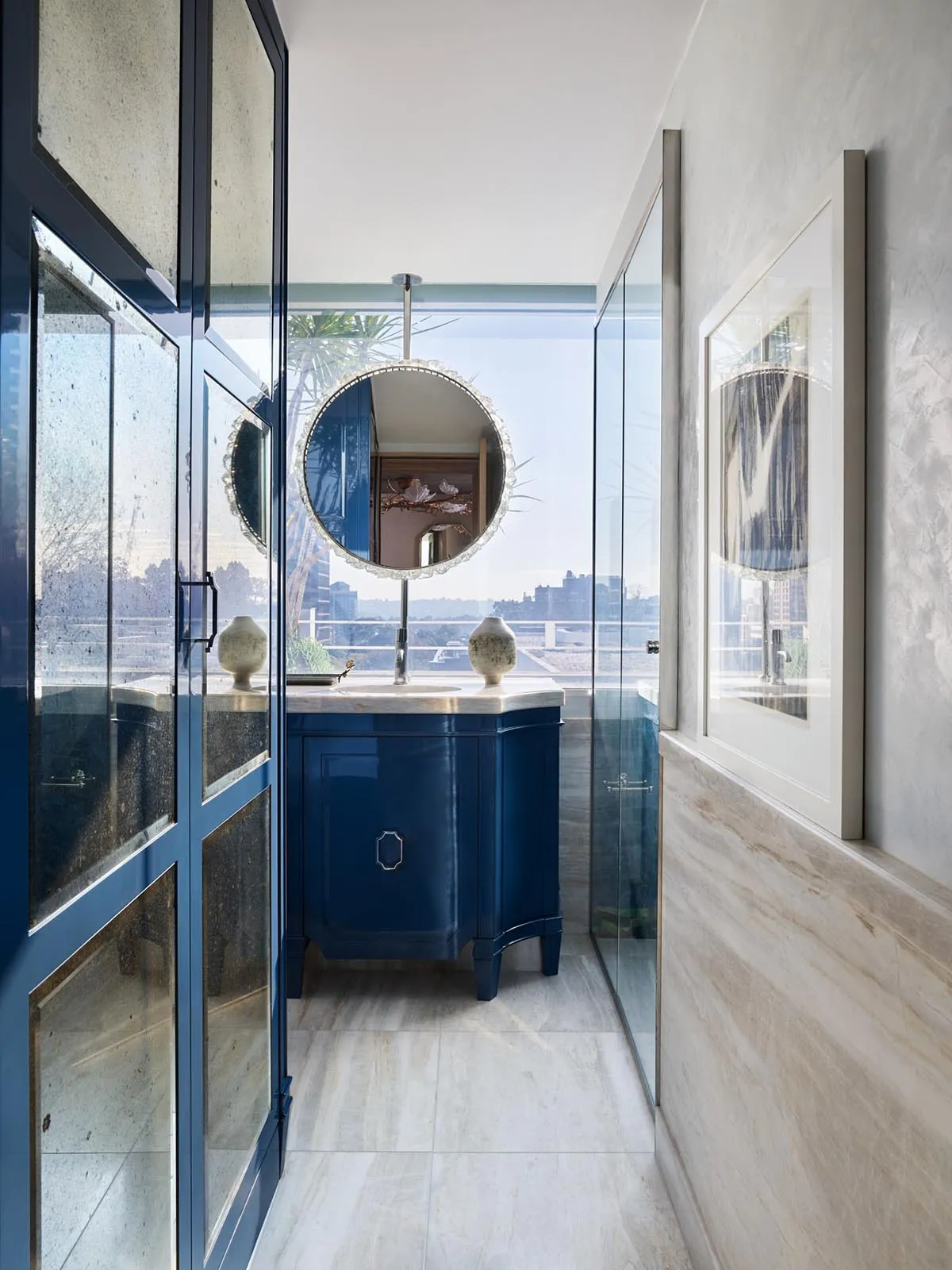 A modern bathroom with blue cabinets, marble countertops, and a circular mirror.