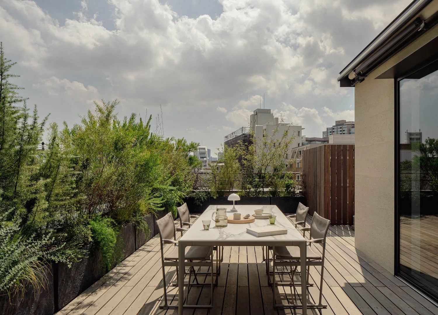 A modern outdoor terrace featuring a concrete dining table and wooden chairs surrounded by lush green plants.