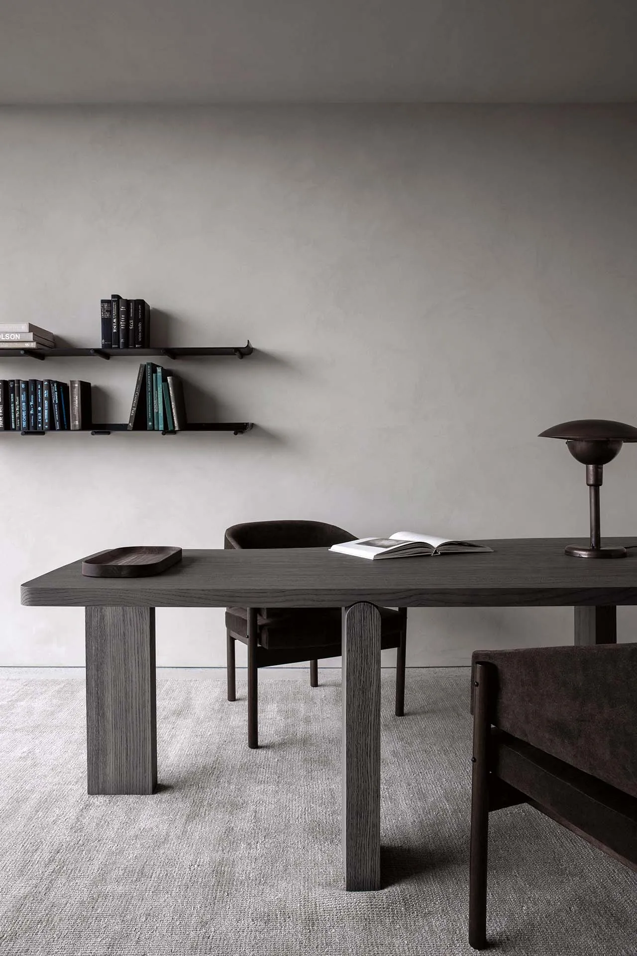 A modern minimalist office space featuring a dark wooden table and a bookshelf filled with books and other items on a gray ca
