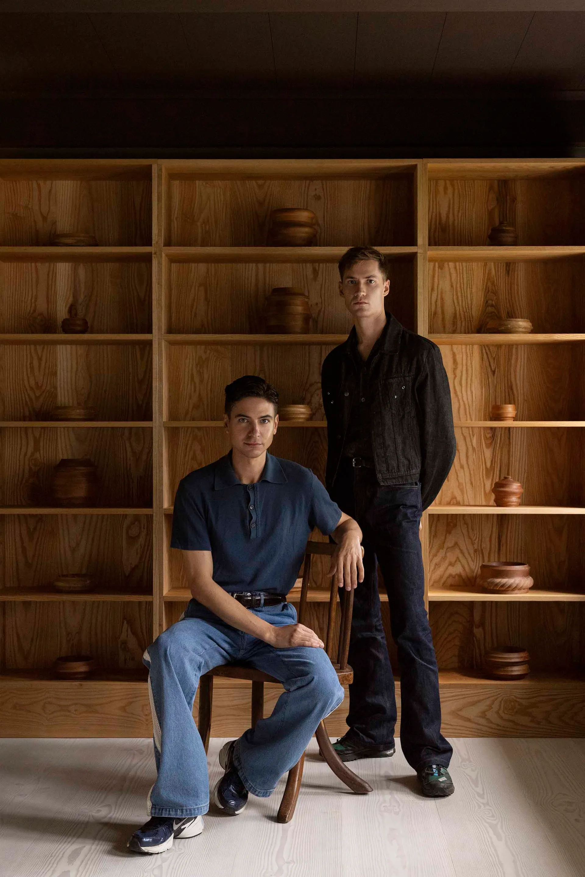 Two men wearing blue jeans standing in a room with wooden shelves.