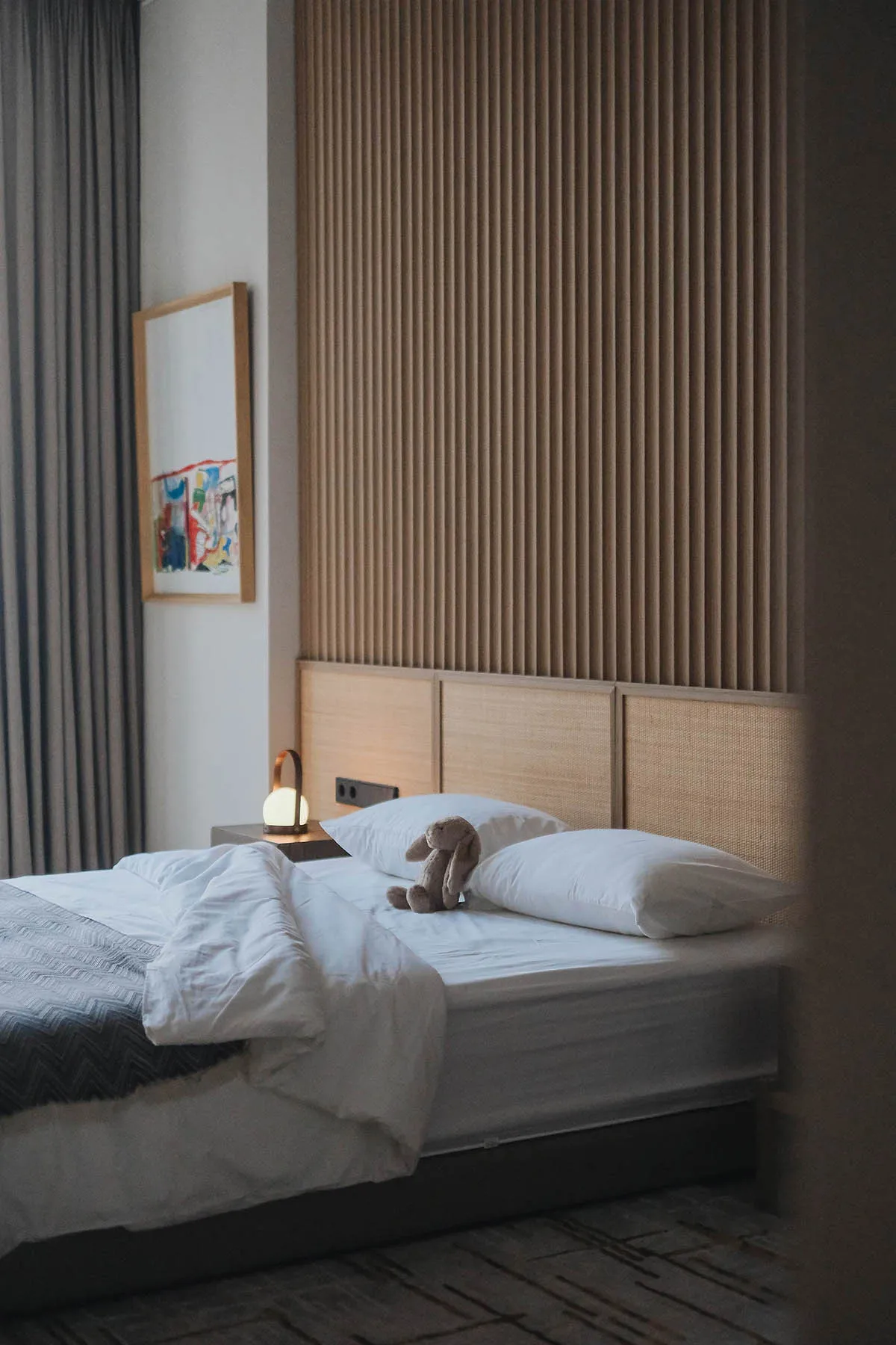 A minimalist bedroom featuring a white wooden headboard, white bedding, and a rabbit plush toy on the bed.