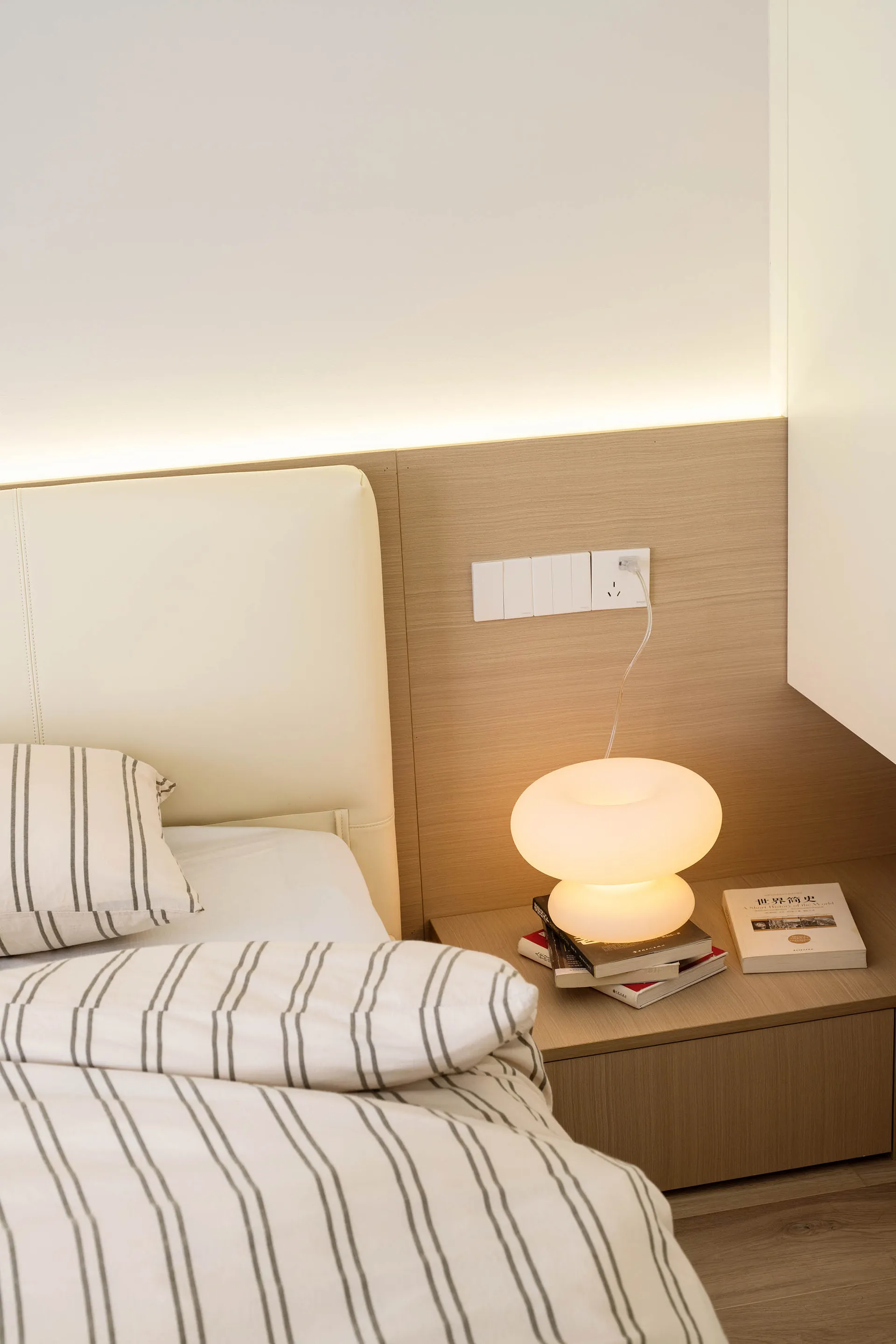 A minimalist bedroom with a white bed, striped bedding, and a wooden headboard and bedside table.