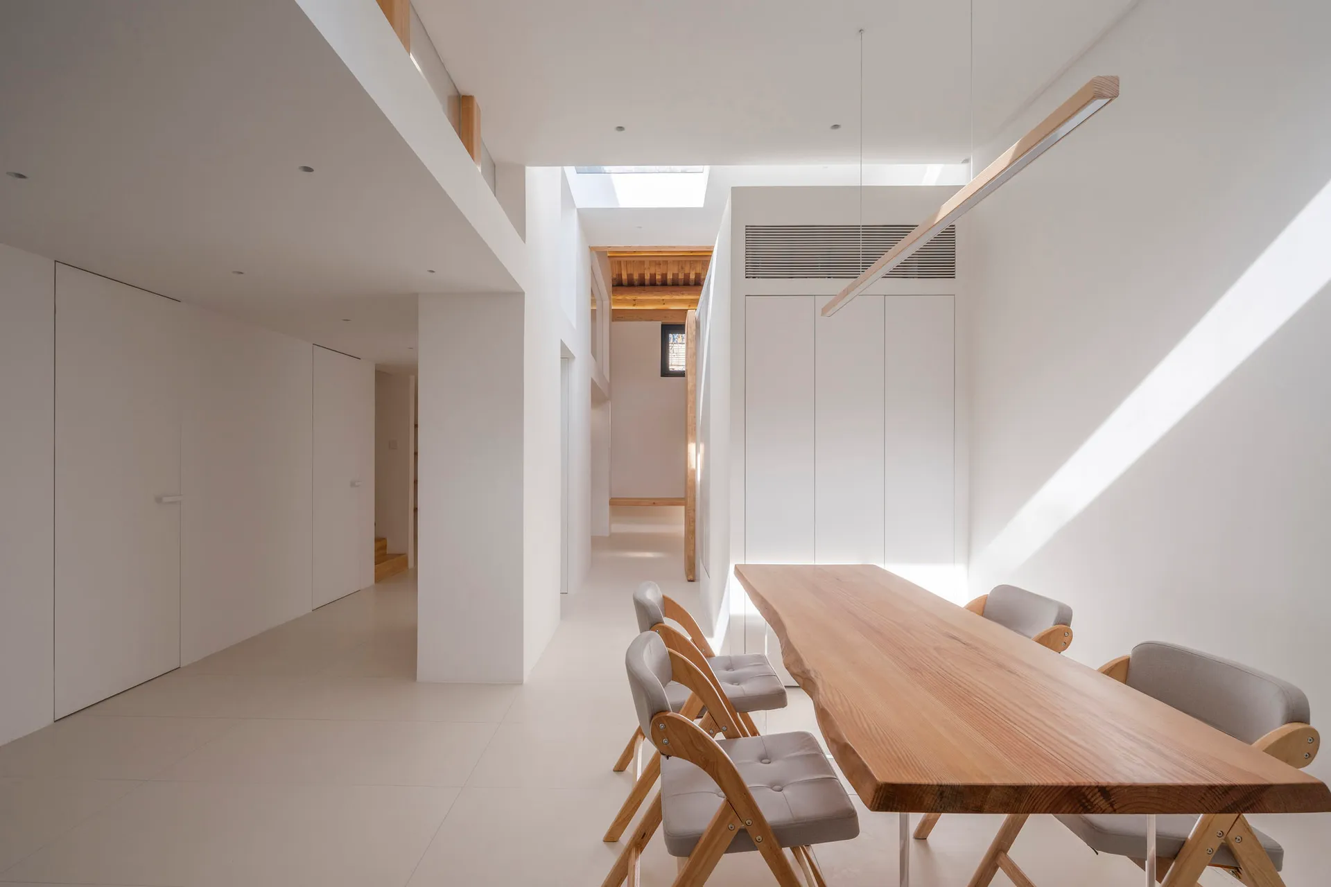 Modern, minimalist dining area with white walls, wooden table, and grey chairs.