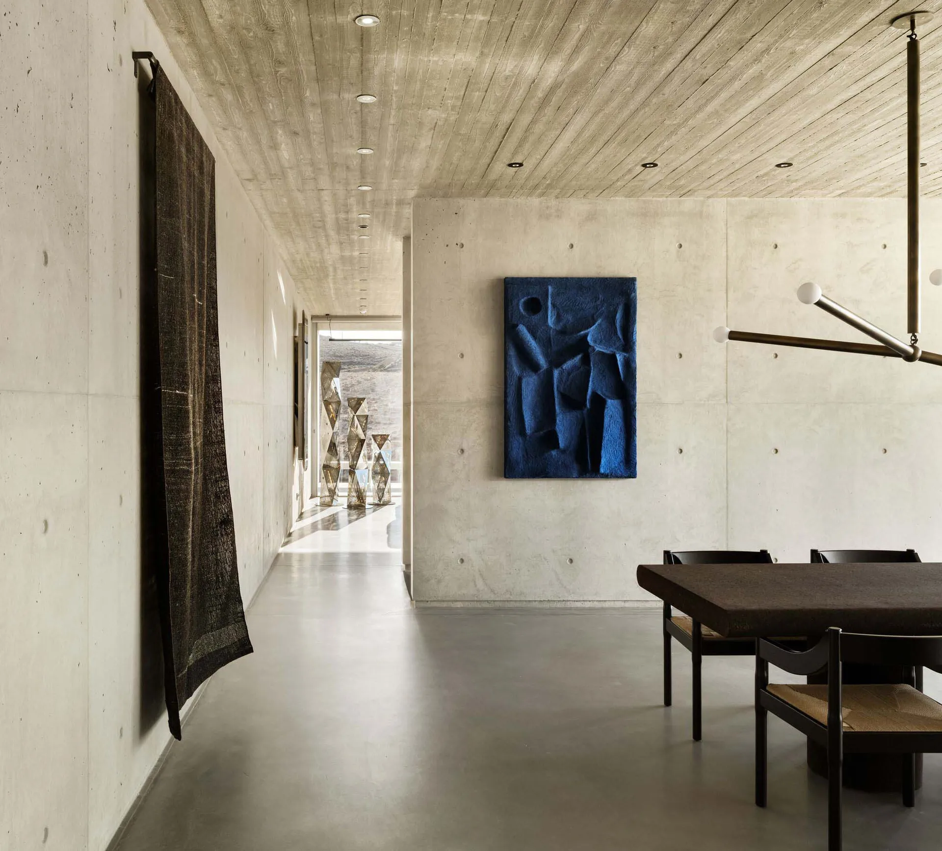 A modern interior space featuring a concrete wall, a blue abstract sculpture, and a wooden dining table with chairs.