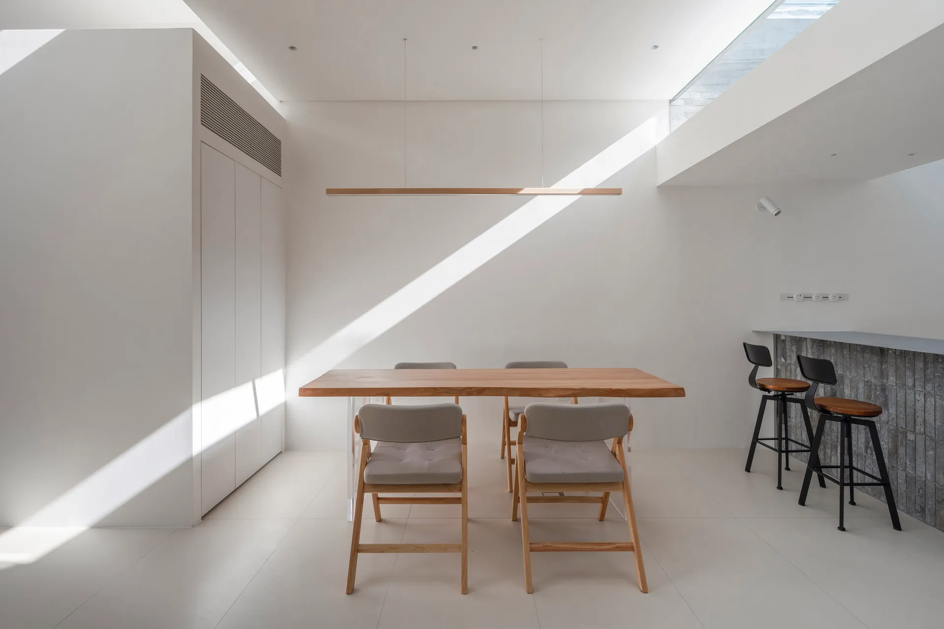 A minimalist dining area with wooden furniture and concrete flooring, featuring a long wooden table and gray upholstered chai