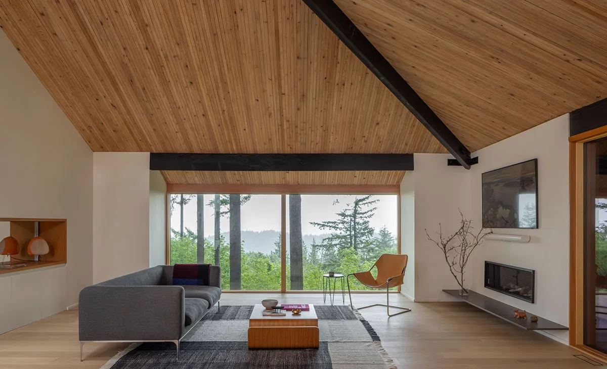 A modern living room with a grey sofa, a wooden coffee table, and a large window overlooking nature.