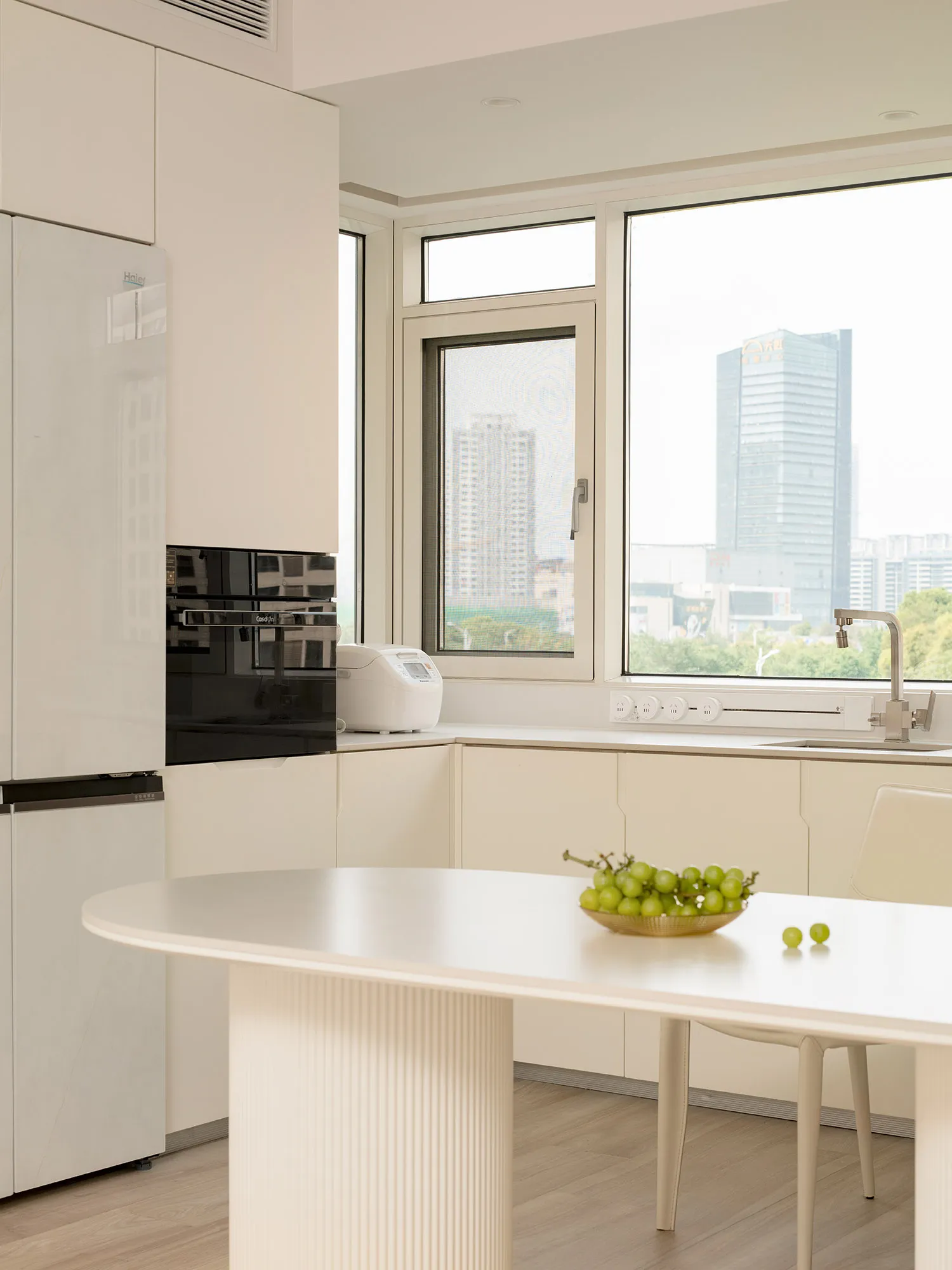 A modern minimalist kitchen featuring white laminate cabinets, a sleek countertop, and a view through a large window.