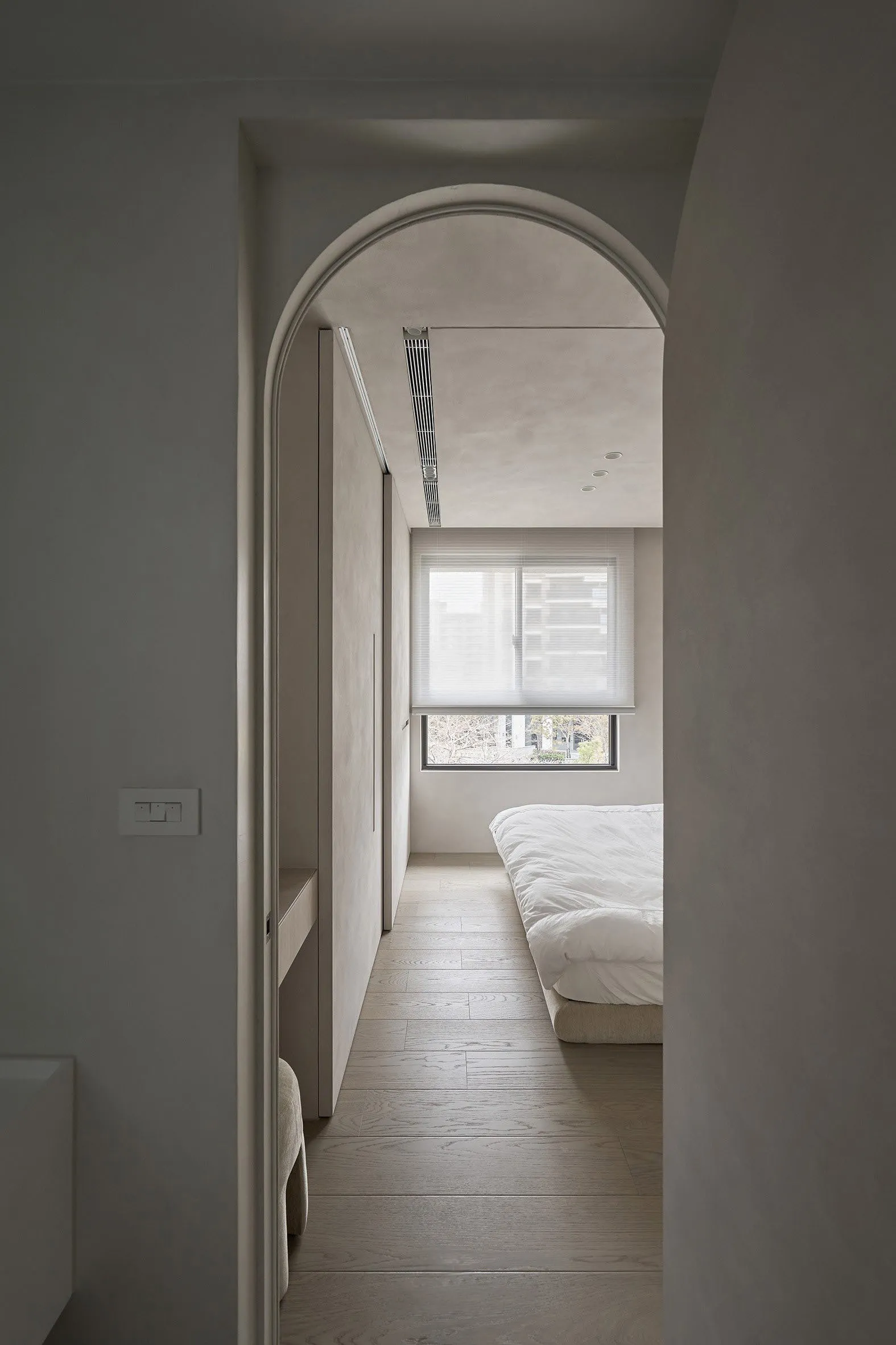 A minimalist bedroom featuring light-colored wood flooring, white bedding, and a large window with blinds.
