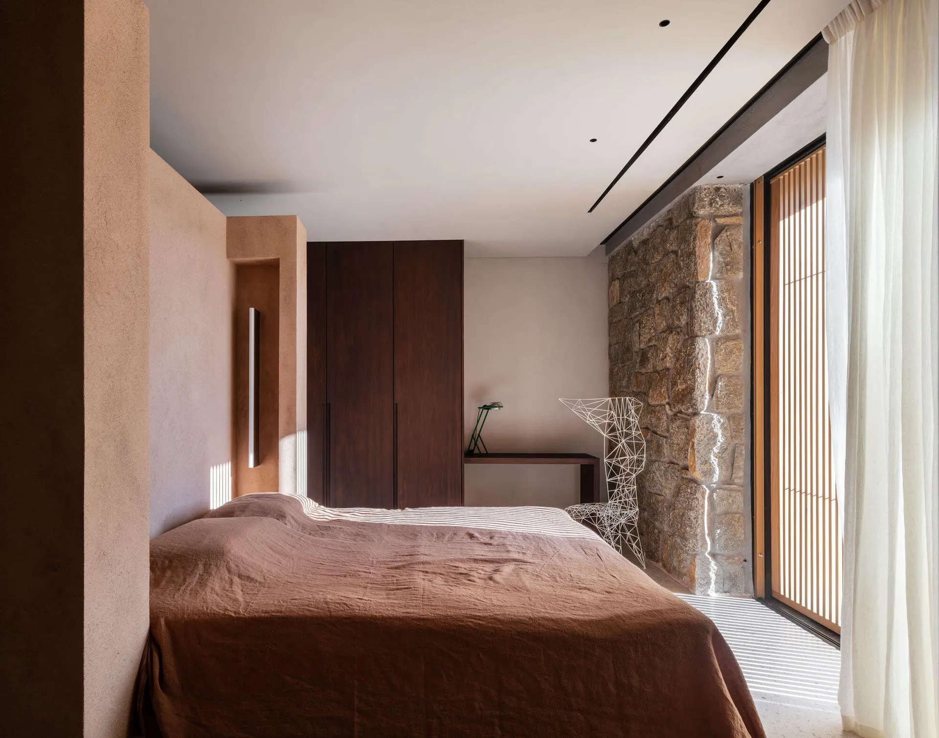 A minimalist bedroom featuring wooden furniture and a stone wall, with natural light streaming through the window.