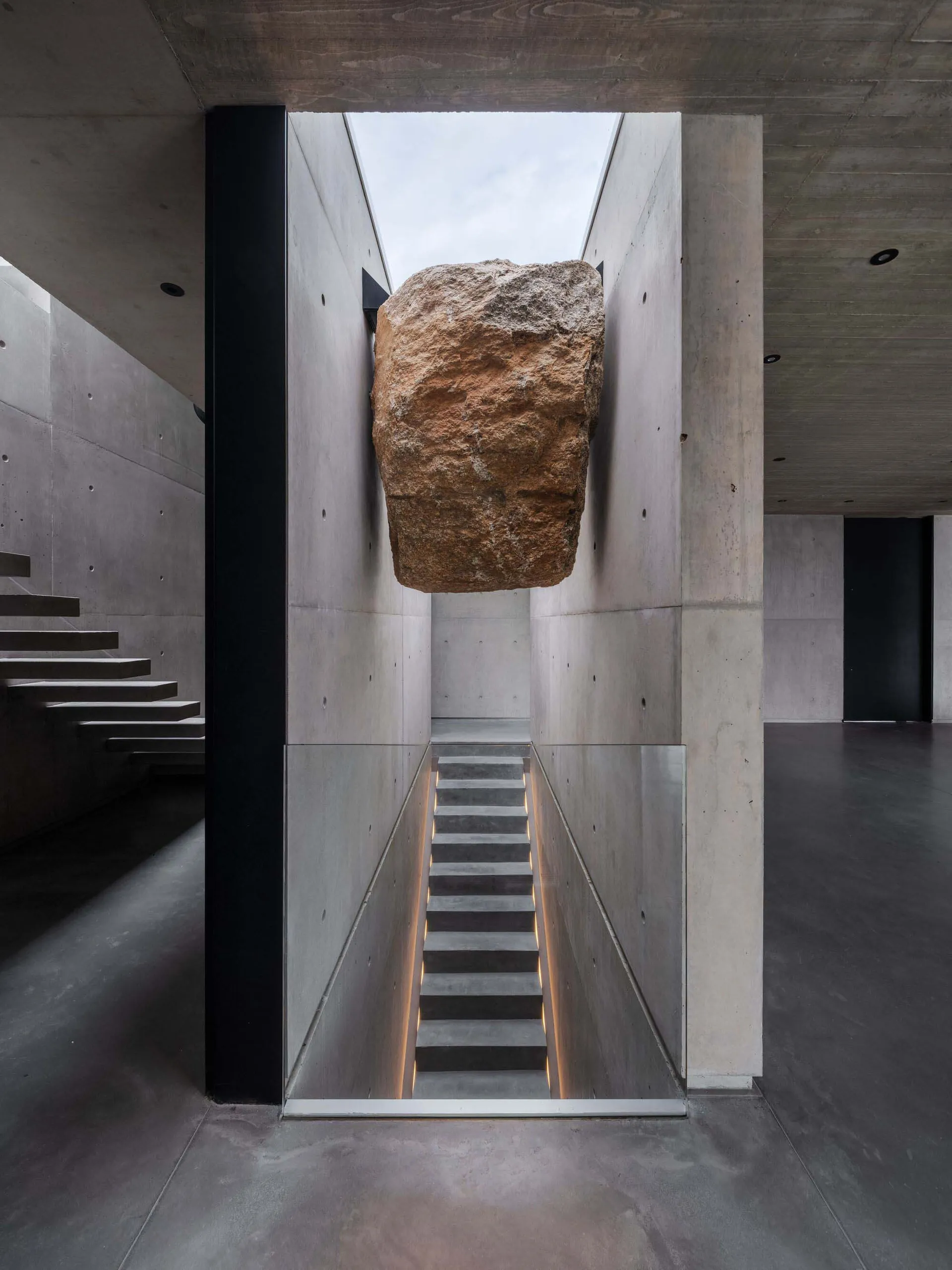 A minimalist stairwell with concrete walls and flooring, featuring a large rock embedded in the ceiling.