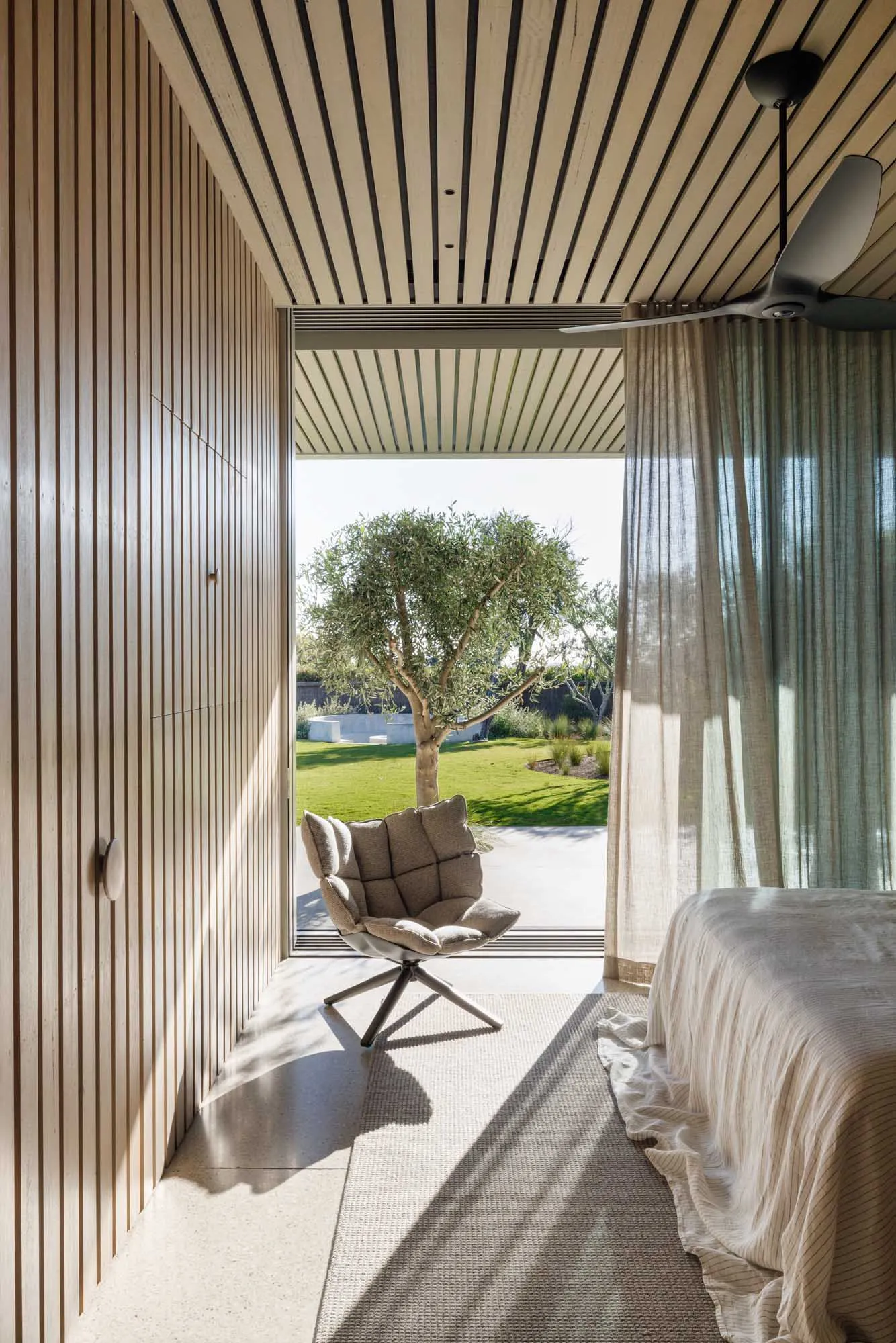 A modern bedroom featuring wooden walls, a cozy chair, and a view of a green tree through a large window.