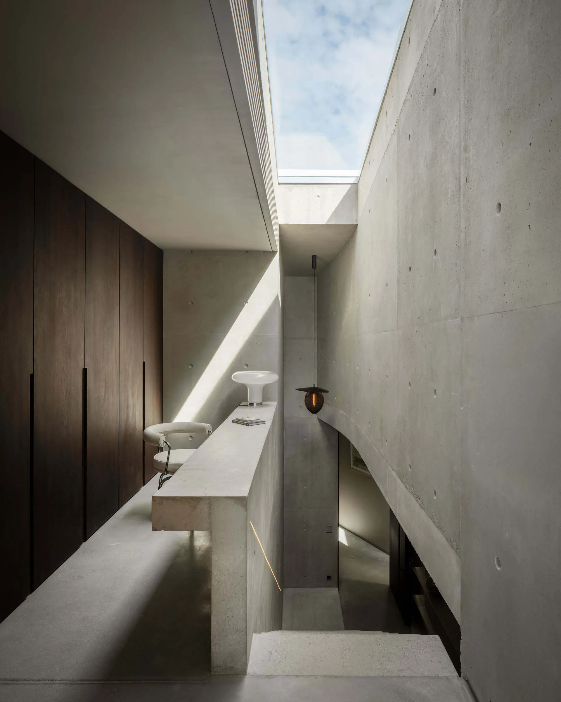 A minimalist corridor with concrete walls, a white chair, and a skylight casting sunlight.
