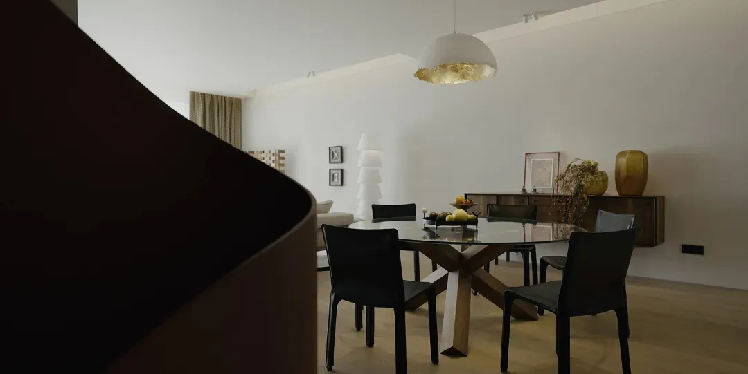 Modern minimalist dining room featuring a round wooden table with glass top and black chairs, illuminated by a minimalist pen