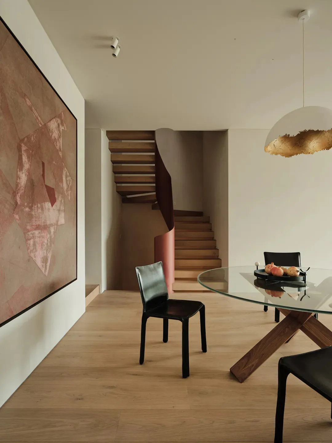 A modern minimalist living room featuring wooden stairs, a glass dining table, and black leather chairs.