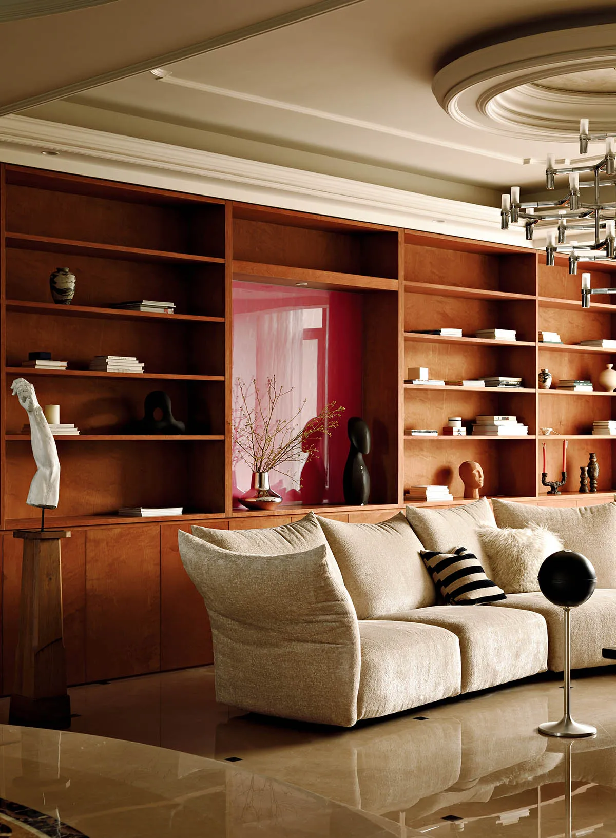 A modern living room with a large beige sofa, wooden shelves filled with books and decorative items, and a marble floor.