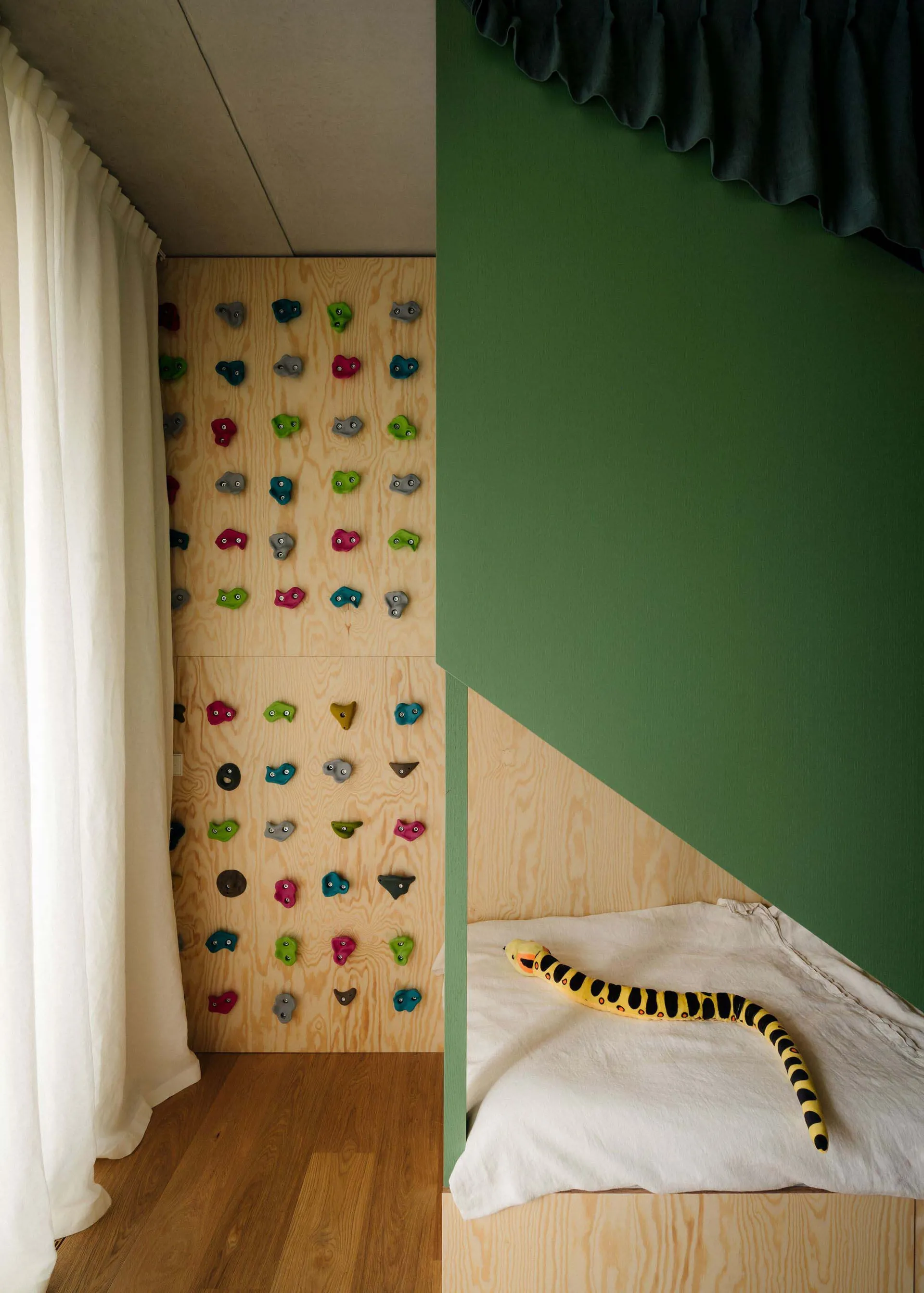 A minimalist playroom featuring a wooden climbing wall with colorful grips and a bed with a snake-shaped plush toy.