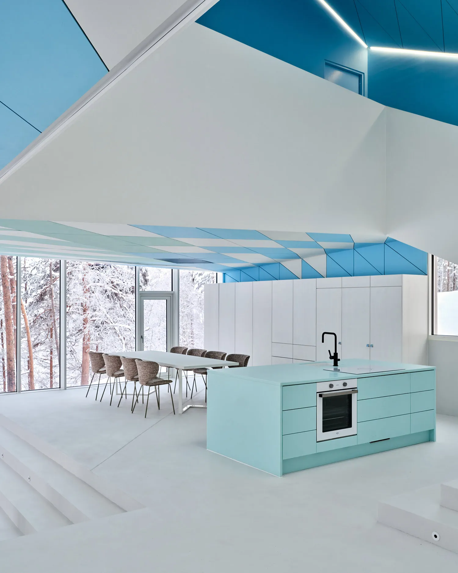 A modern kitchen with a turquoise island, white countertops, and a long dining table surrounded by chairs, overlooking a snow