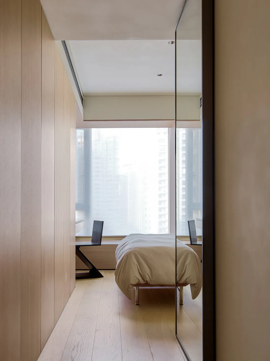 Modern minimalist bedroom with light wood floors, glass window overlooking cityscape, and metal furniture.