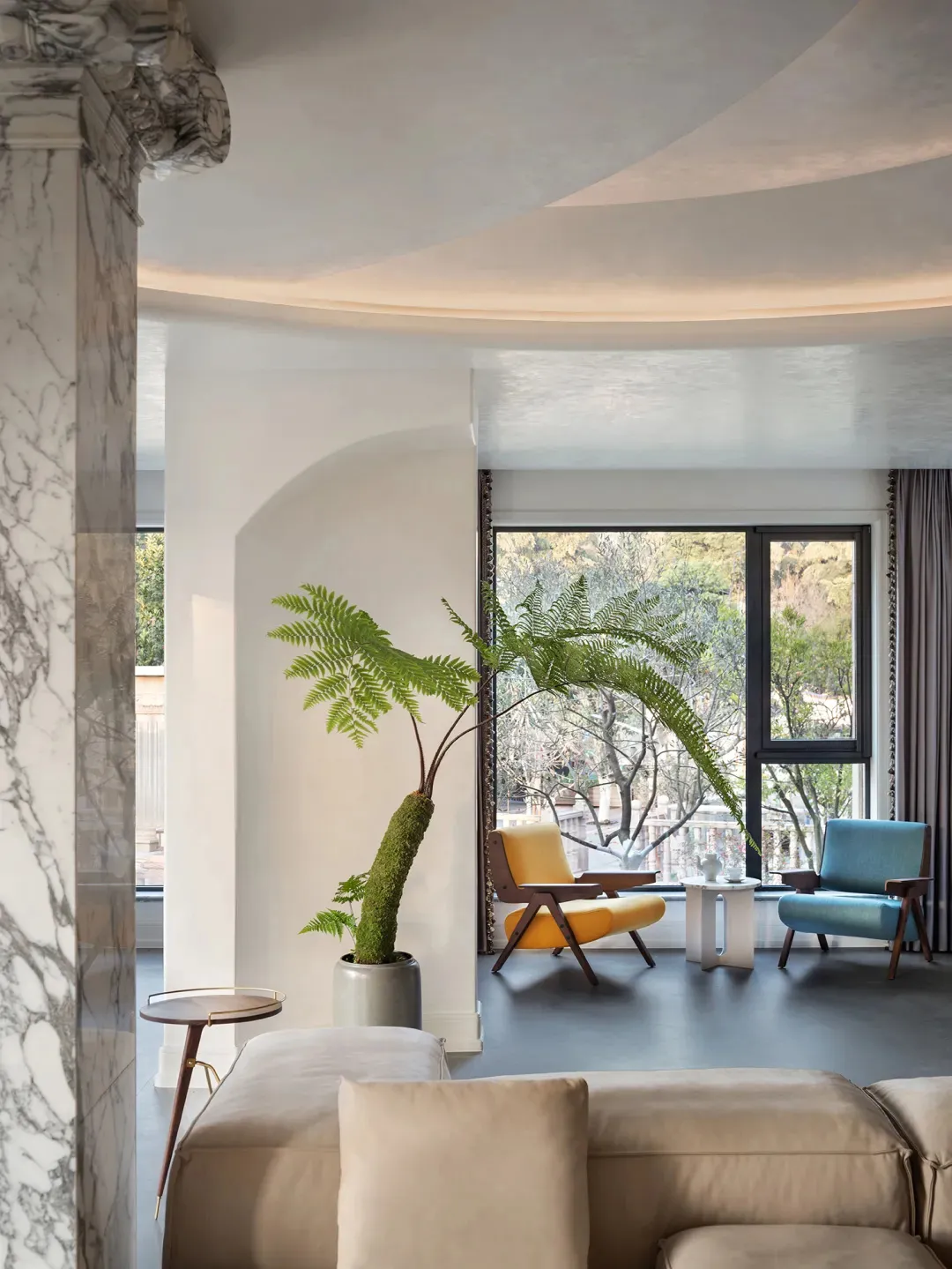 A modern minimalist living room with concrete walls, a beige sofa, a yellow chair, a blue chair, and a potted fern plant.