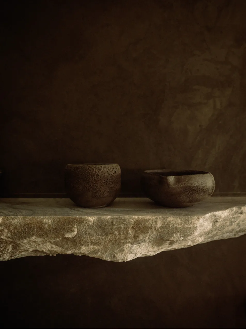 Two ceramic bowls made of clay material, placed on a stone shelf against a dark brown wall.
