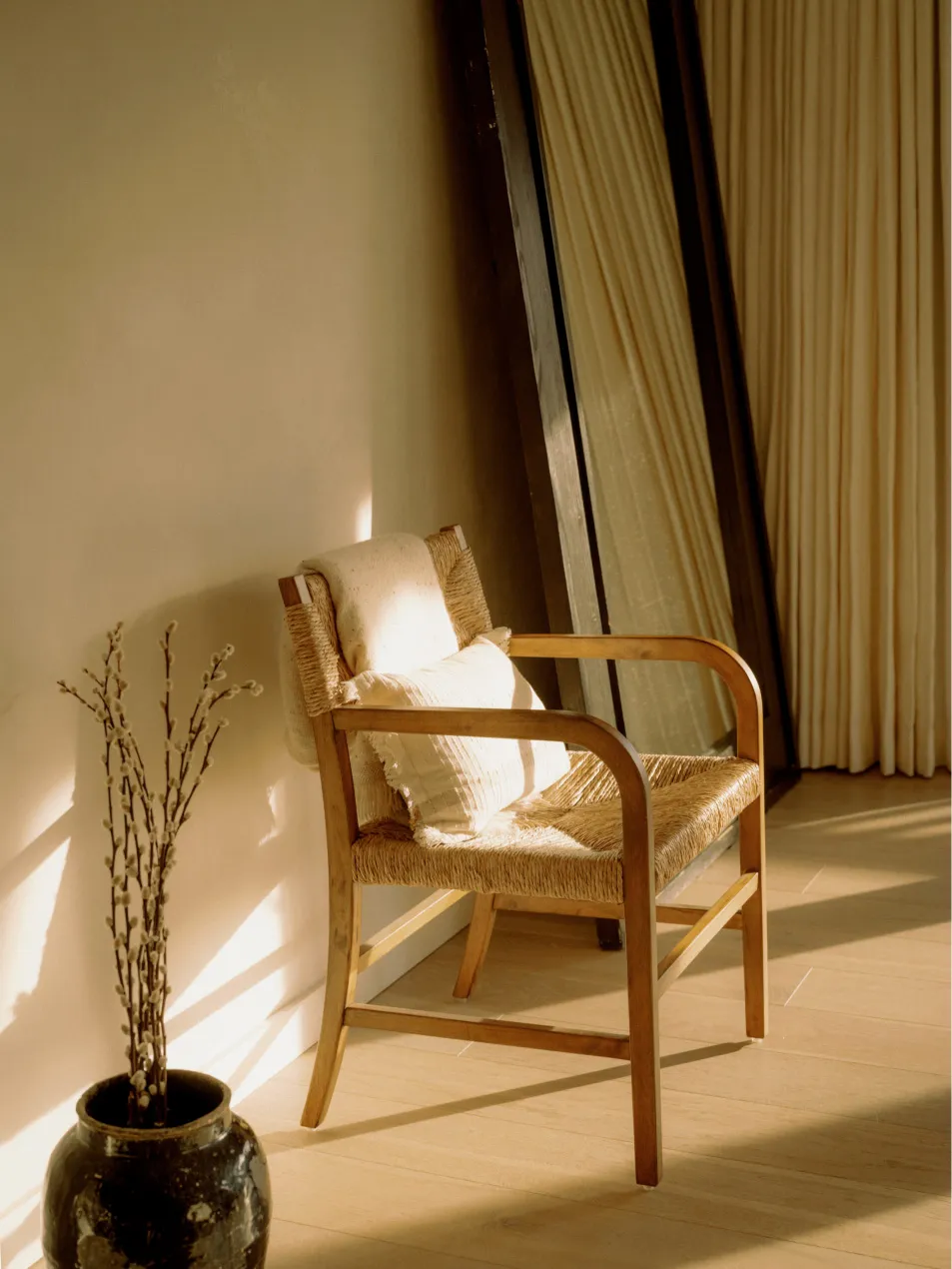 A minimalist living room with a wooden armchair and cream-colored curtains, featuring sunlight streaming through a window.