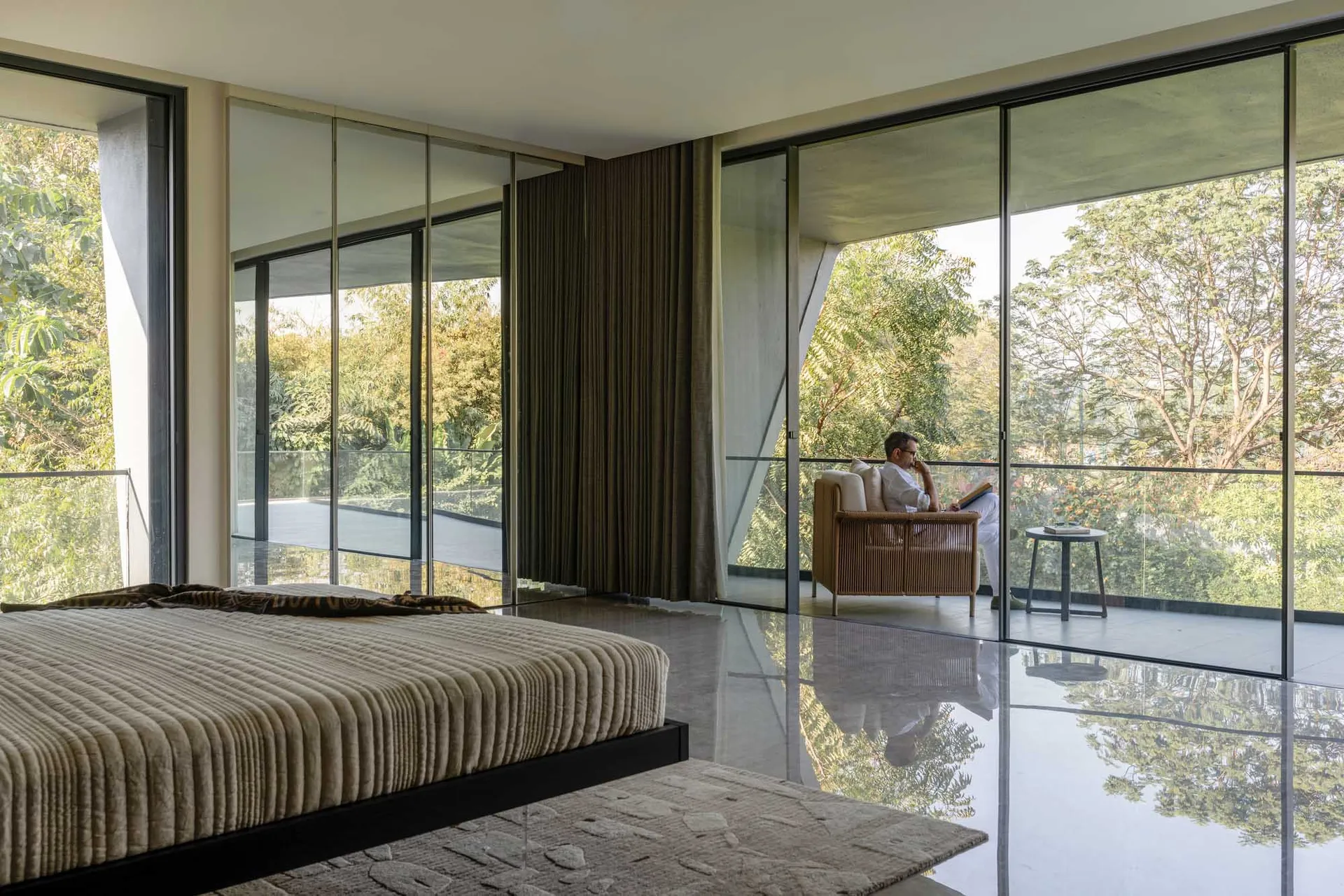 A modern residential casual living room with a large bed, a person reading in a sofa chair on the terrace, and a view of lush