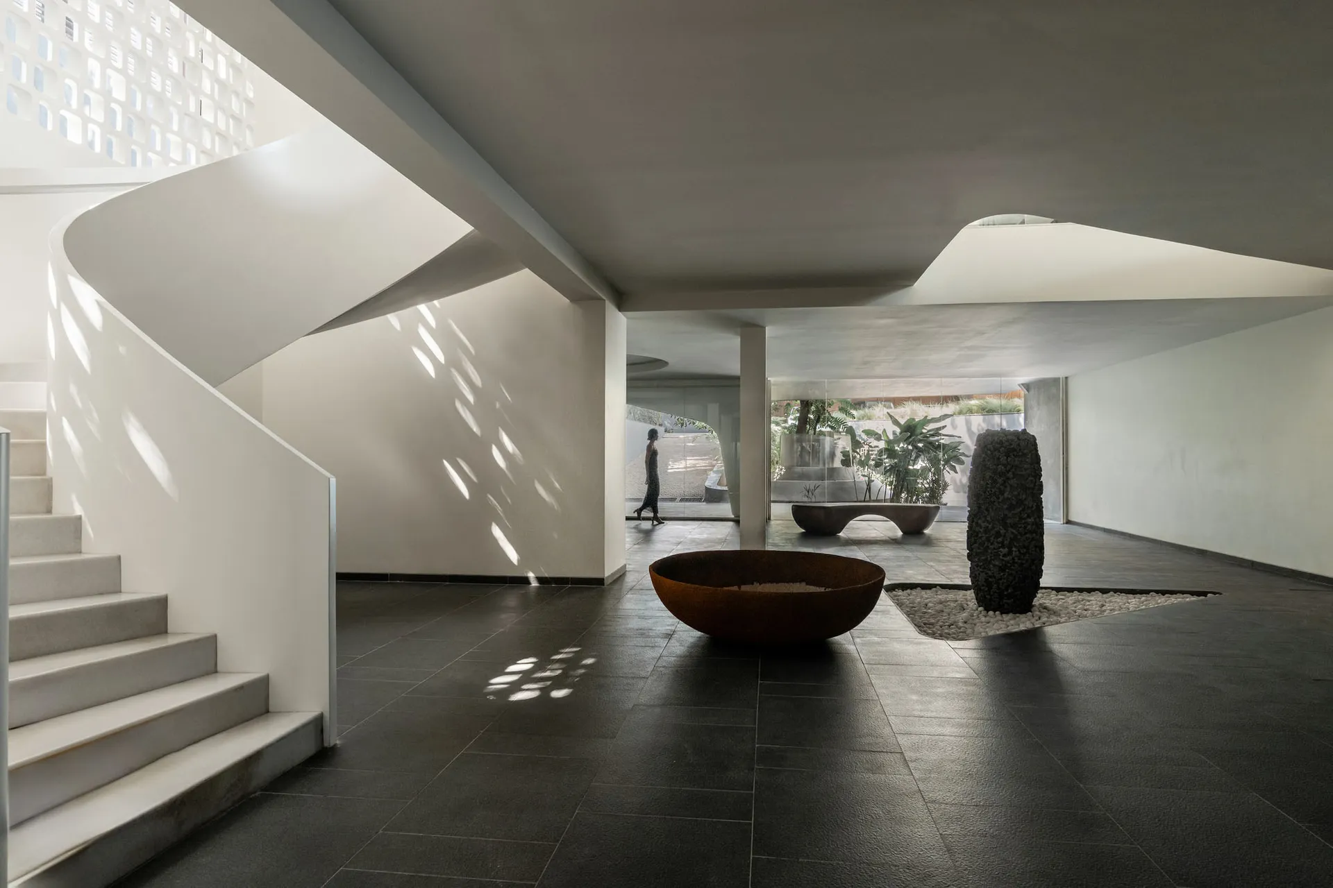 Modern lounge area with concrete walls and floor, featuring a spiral staircase, a large round metal planter, and greenery in 