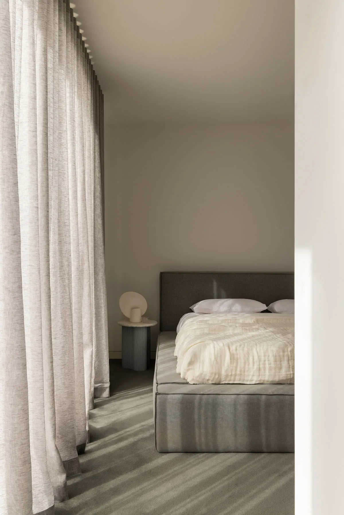 A modern bedroom featuring a grey upholstered bed with white bedding, surrounded by sheer curtains and a textured grey carpet