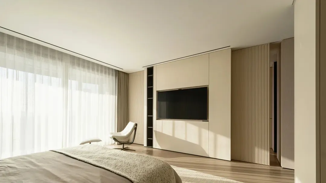 A minimalist bedroom with a large window, white bedding, and a white chair next to a large wooden cabinet.