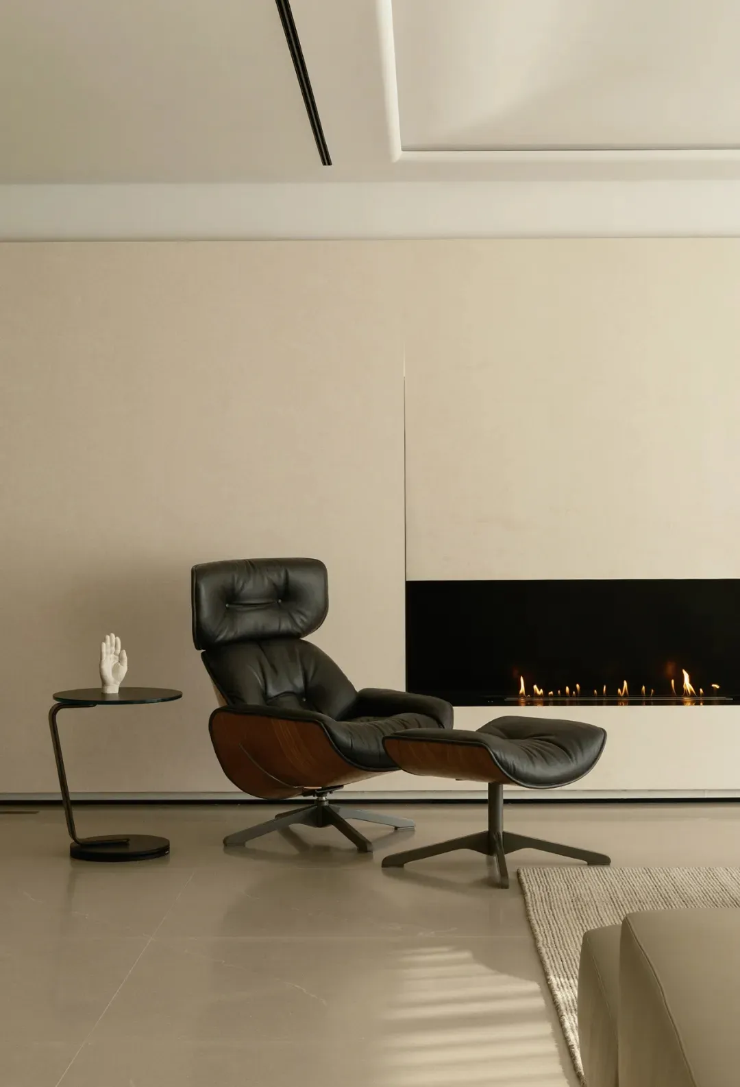 A minimalist living room featuring a black leather and wood armchair with a footstool, a side table with a sculpture, and a m