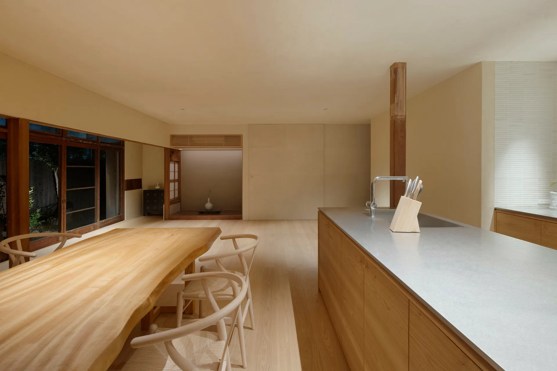 A minimalist residential kitchen with wooden countertops, flooring, and chairs, featuring a large kitchen island with a sink.