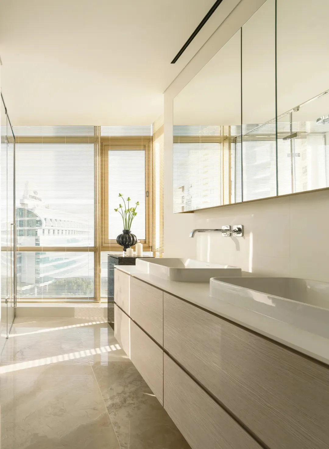 A modern bathroom with white marble flooring, wooden cabinets, and a large window with bamboo blinds.