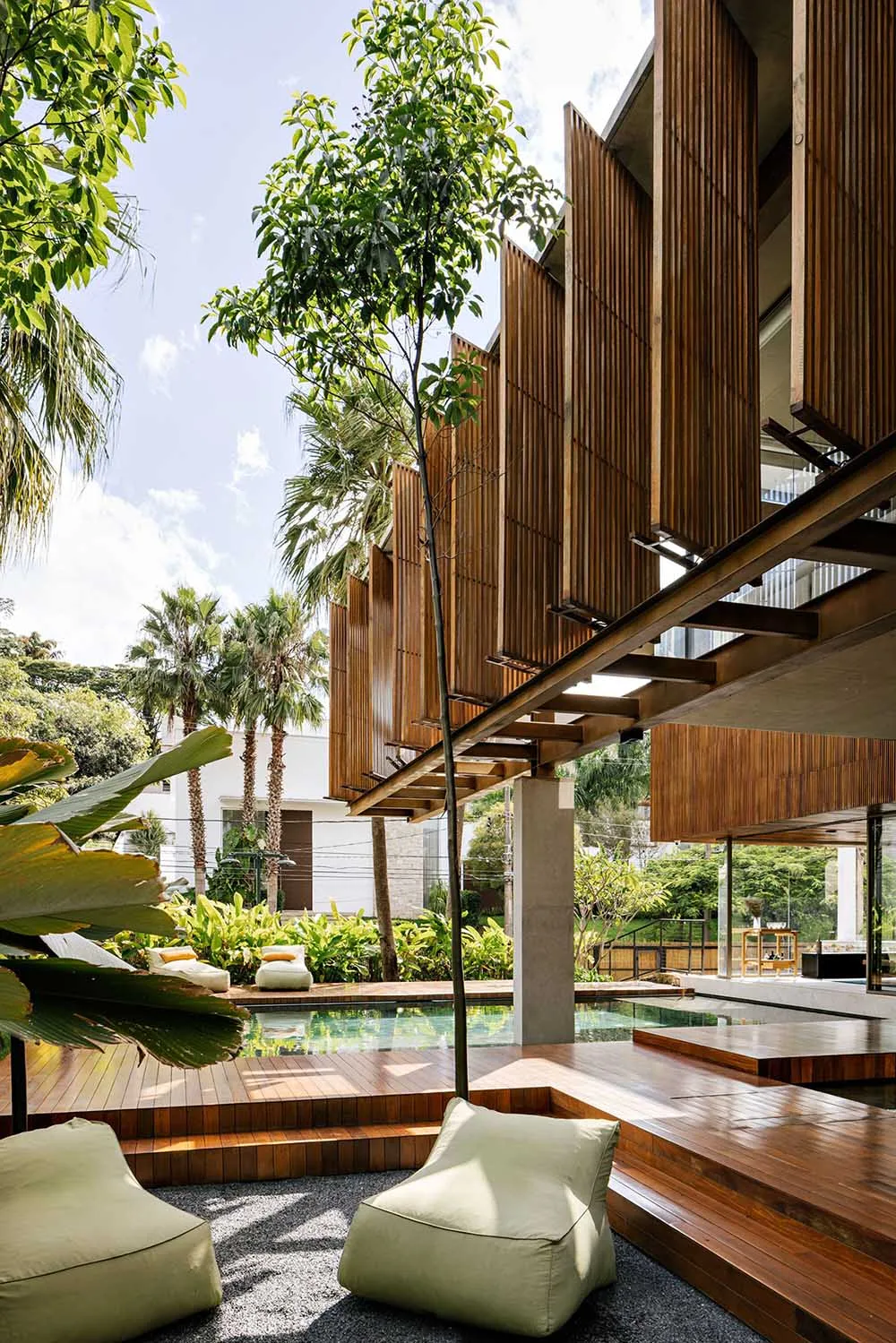 A modern residential exterior featuring wooden panels and a large swimming pool surrounded by greenery.