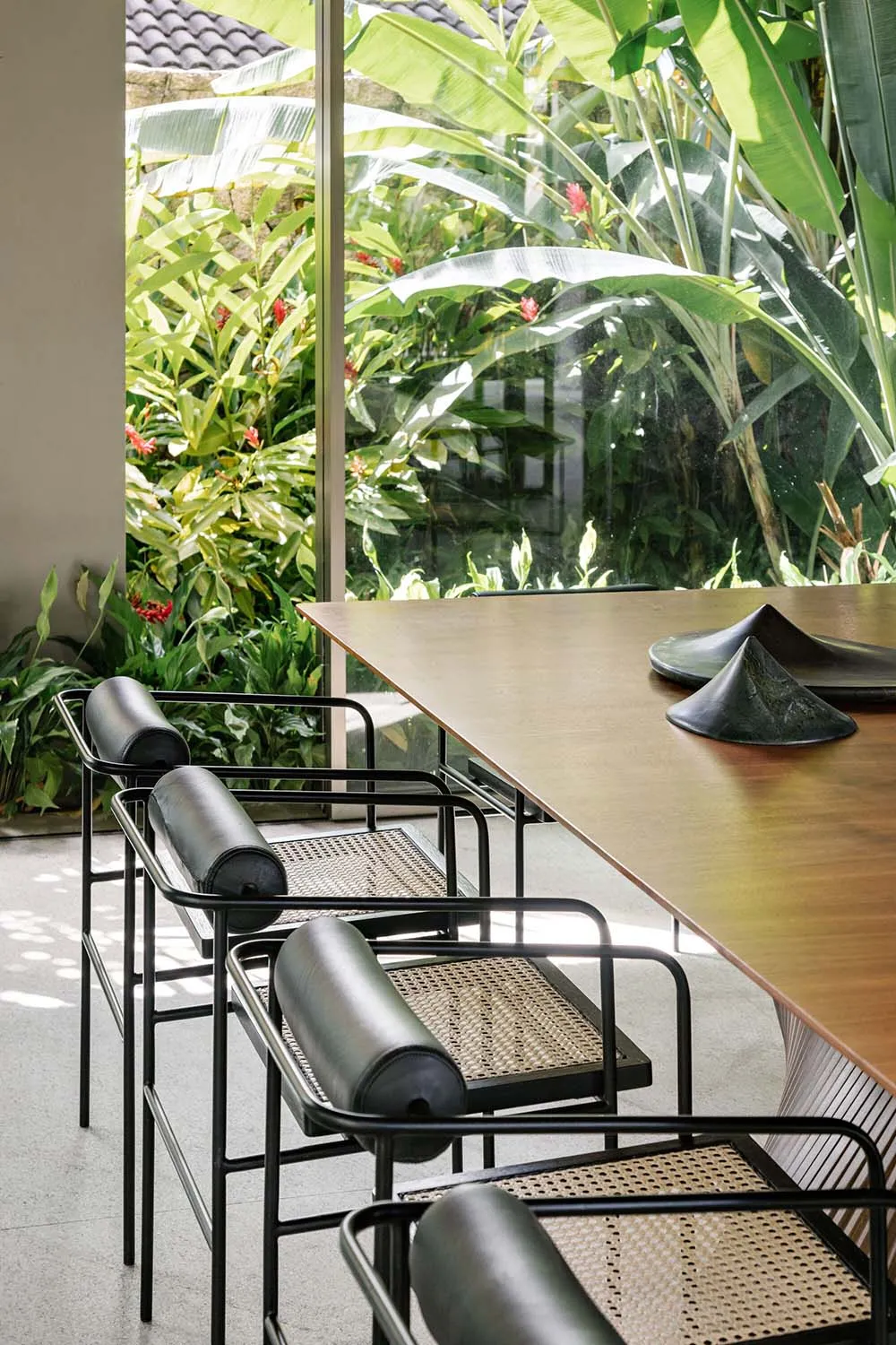Modern minimalist dining table with black metal frame and woven rattan seating surrounded by lush greenery through large wind