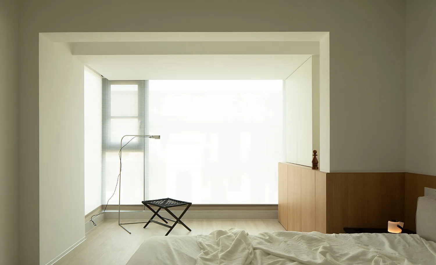 A modern minimalist bedroom with a large window, natural light, a bed, and a black ottoman.