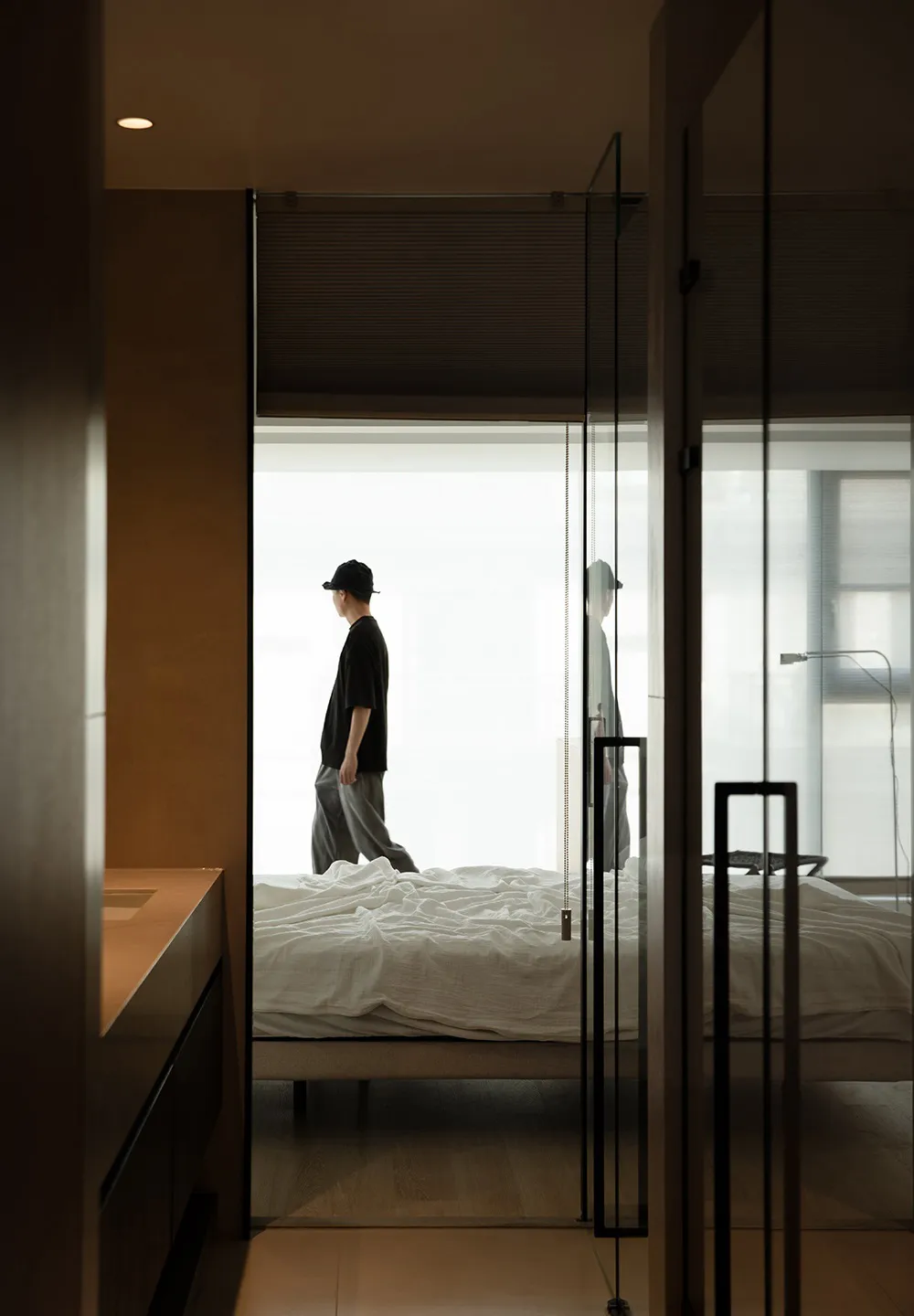 A minimalist bedroom with a bed covered in white linen, natural light streaming in through the window, and a person walking i