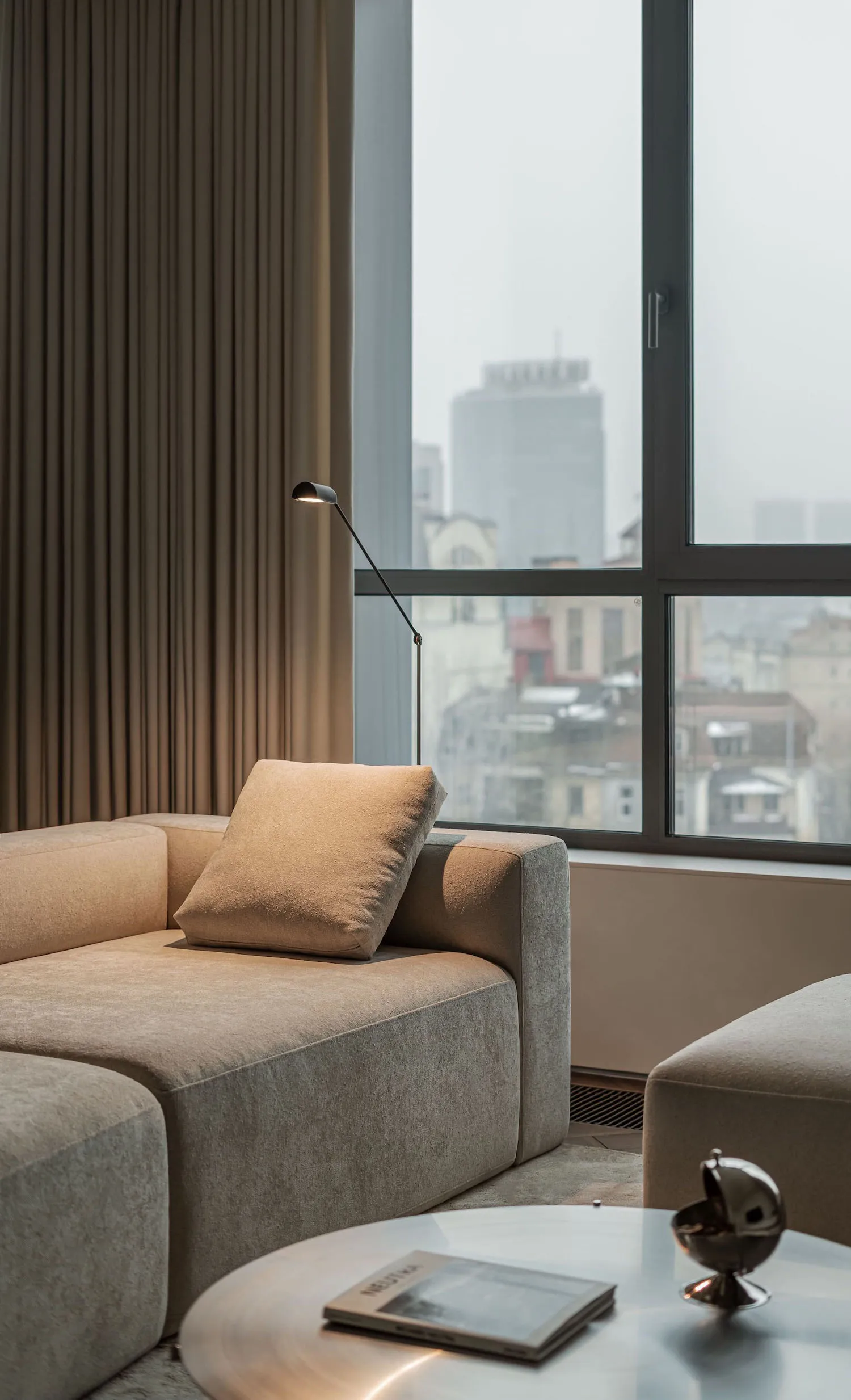 Modern minimalist living room featuring a comfortable beige fabric sofa, a sleek table lamp, and a cityscape view through lar