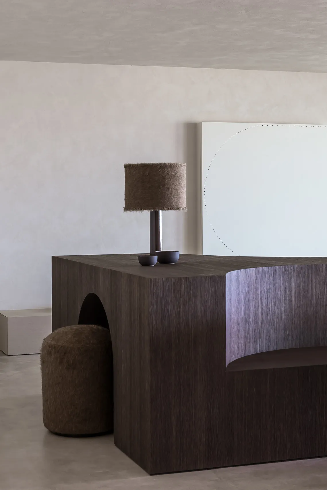 A modern interior design featuring a wooden table with a lamp, a white wall with a circular pattern, and a plush ottoman.