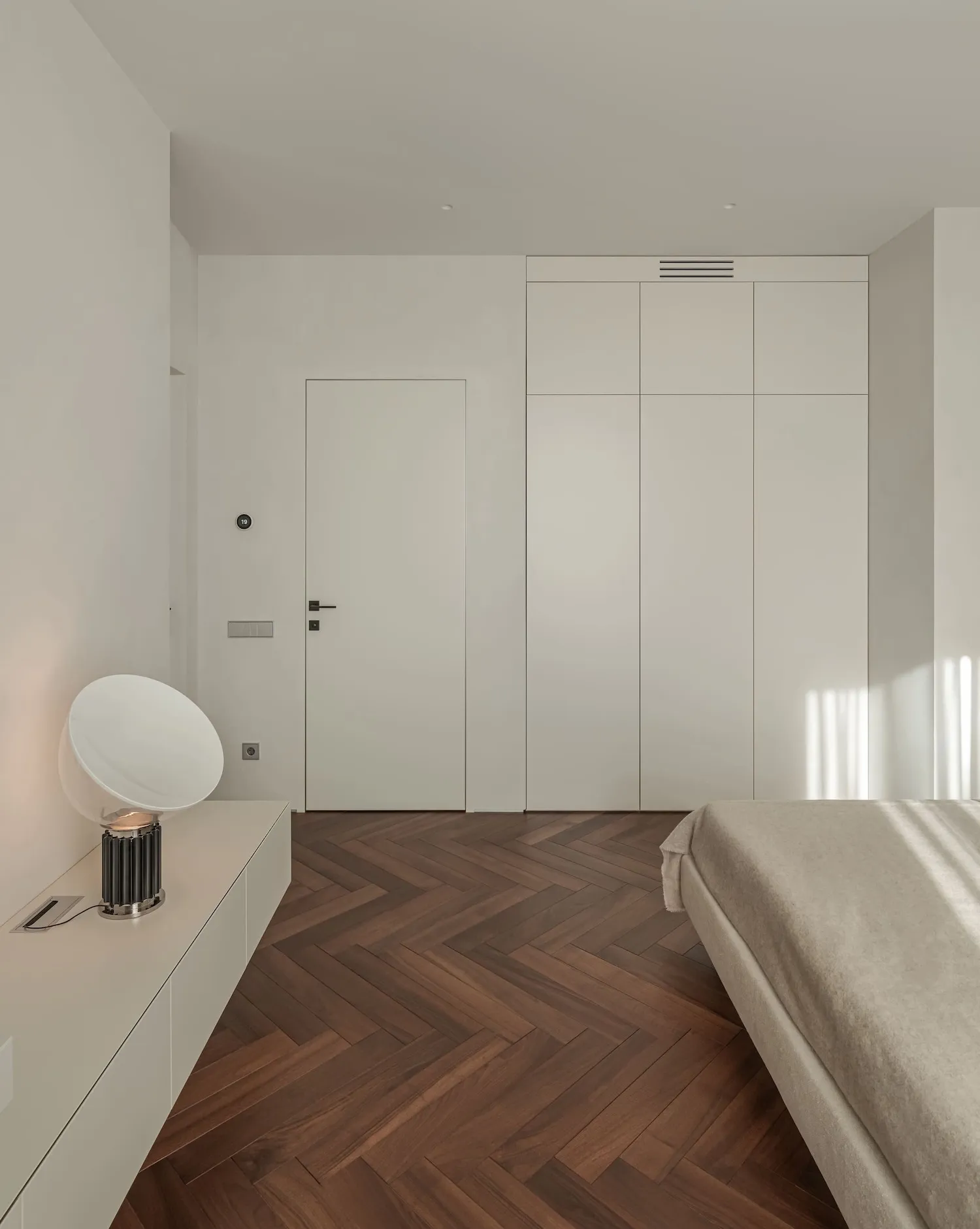 Modern minimalist bedroom with white walls, wooden flooring, and a single white lamp on a floating nightstand.
