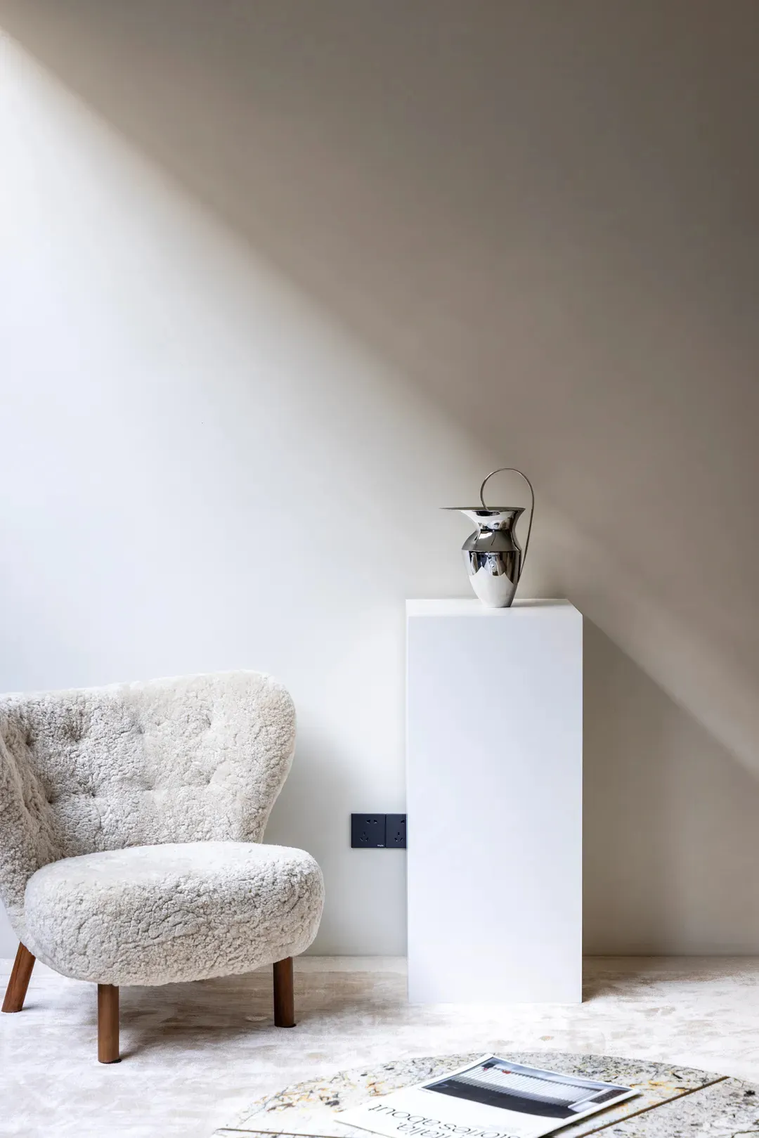A minimalist living room with a sheepskin armchair, a metallic vase on a pedestal, and an open book on a stone table with a m