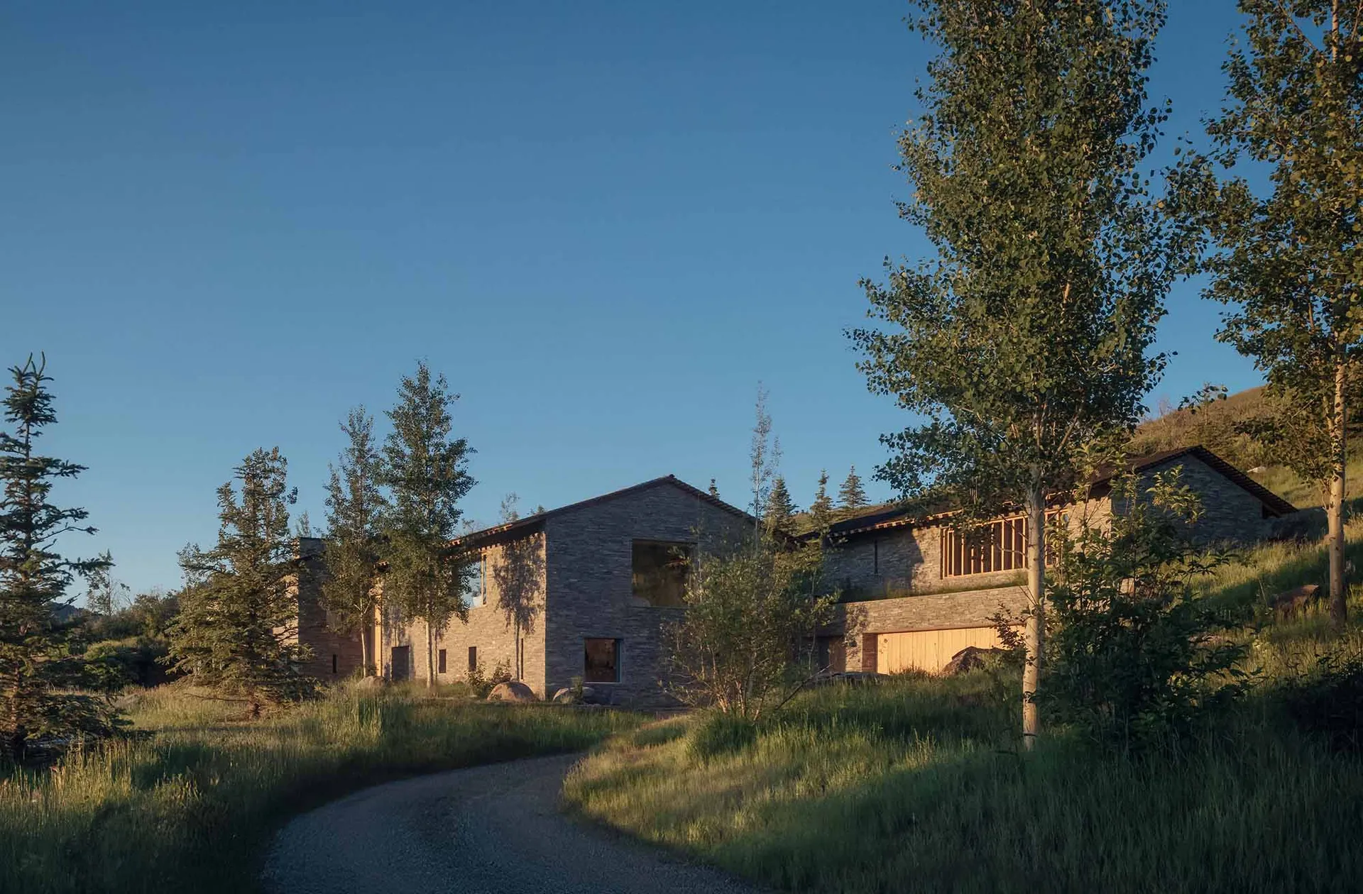 A modern residential house with stone and wood exterior surrounded by greenery and dirt road.