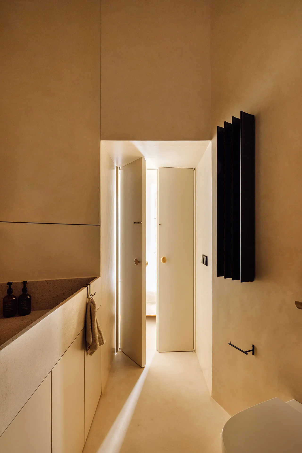 A minimalist bathroom with concrete walls and floor, featuring a sink and a toilet with a black radiator and a doorway leadin