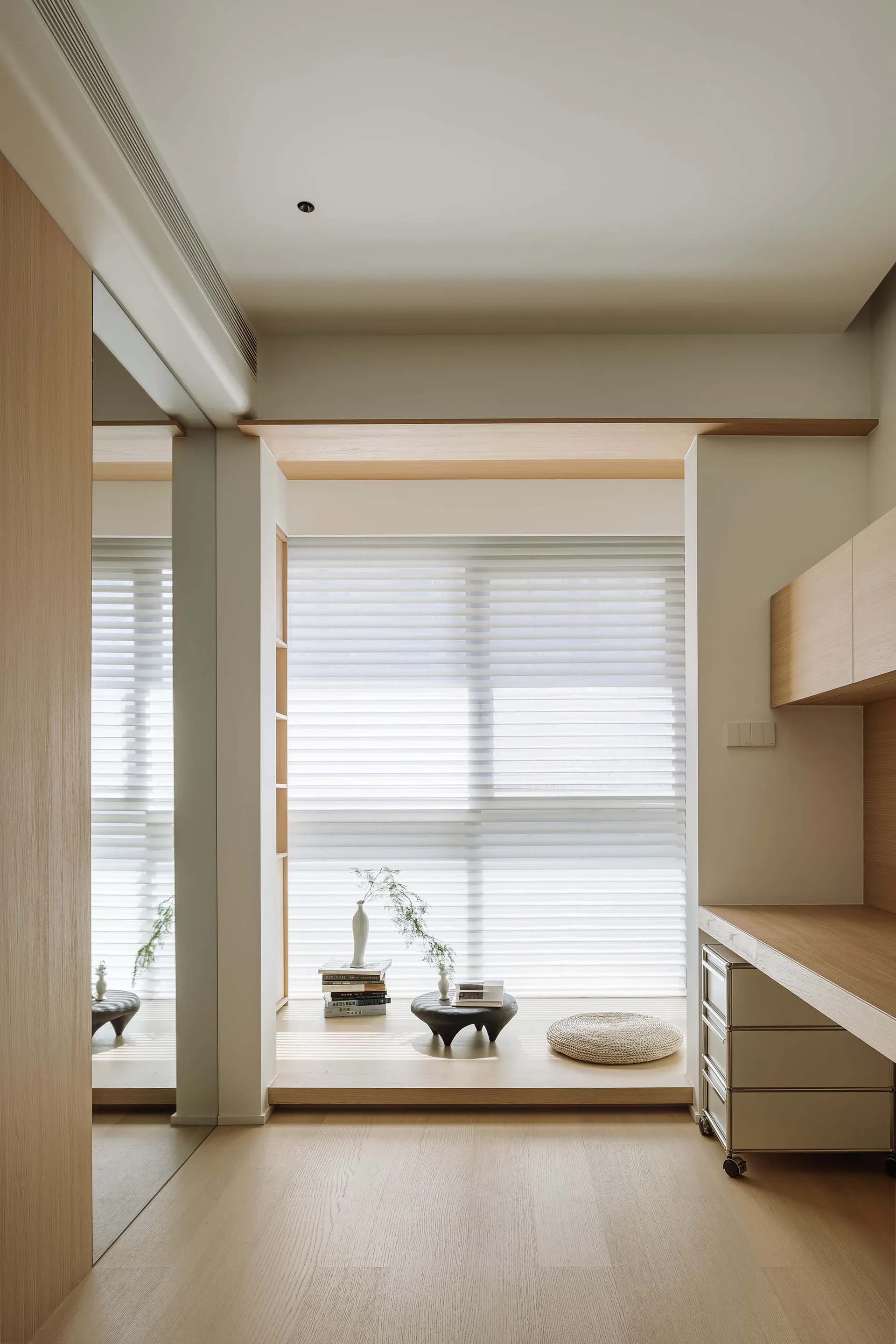 Minimalist residential interior featuring wooden floors, glass doors, and a concrete bench by a window with a vase and stack