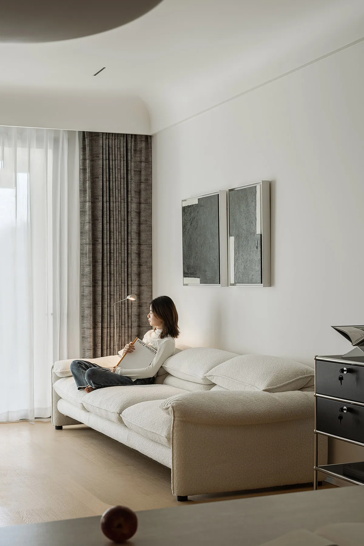 A modern minimalist living room features a white couch with plush cushions and a large window with sheer drapes, allowing nat