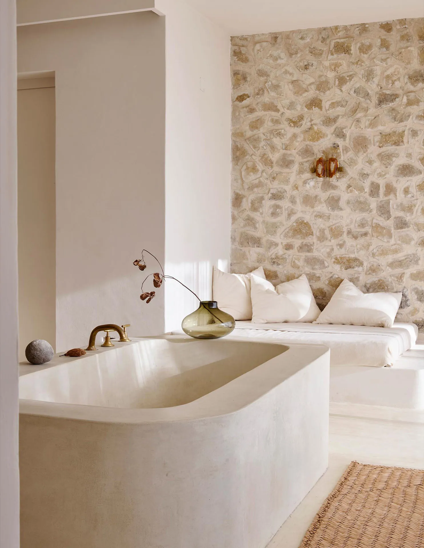 A minimalist bathroom featuring a concrete tub and a cozy nook with a stone wall and white cushions.