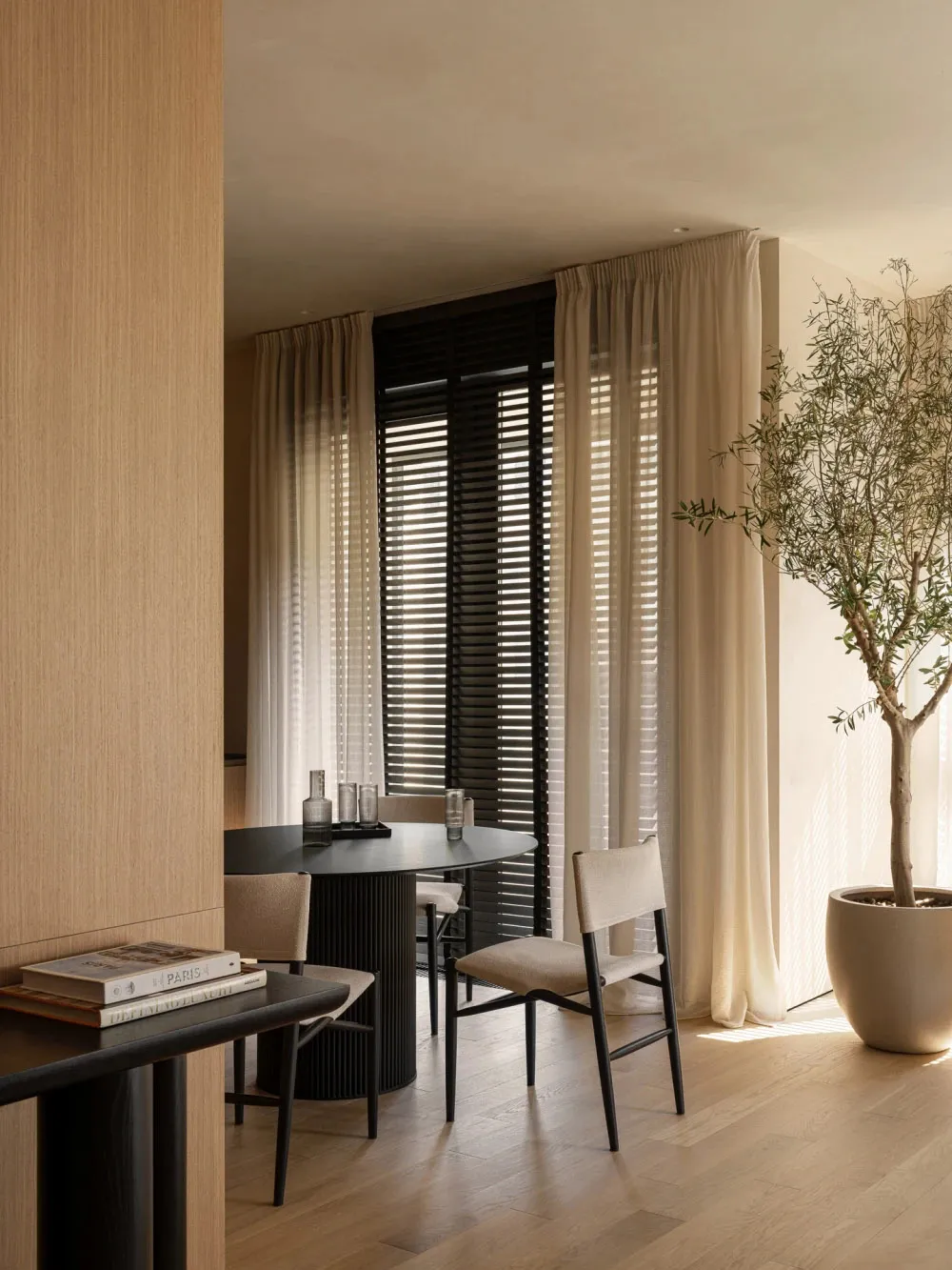 A modern interior design featuring wooden elements and lighting. The space includes a round table, chairs, and a tall plant.