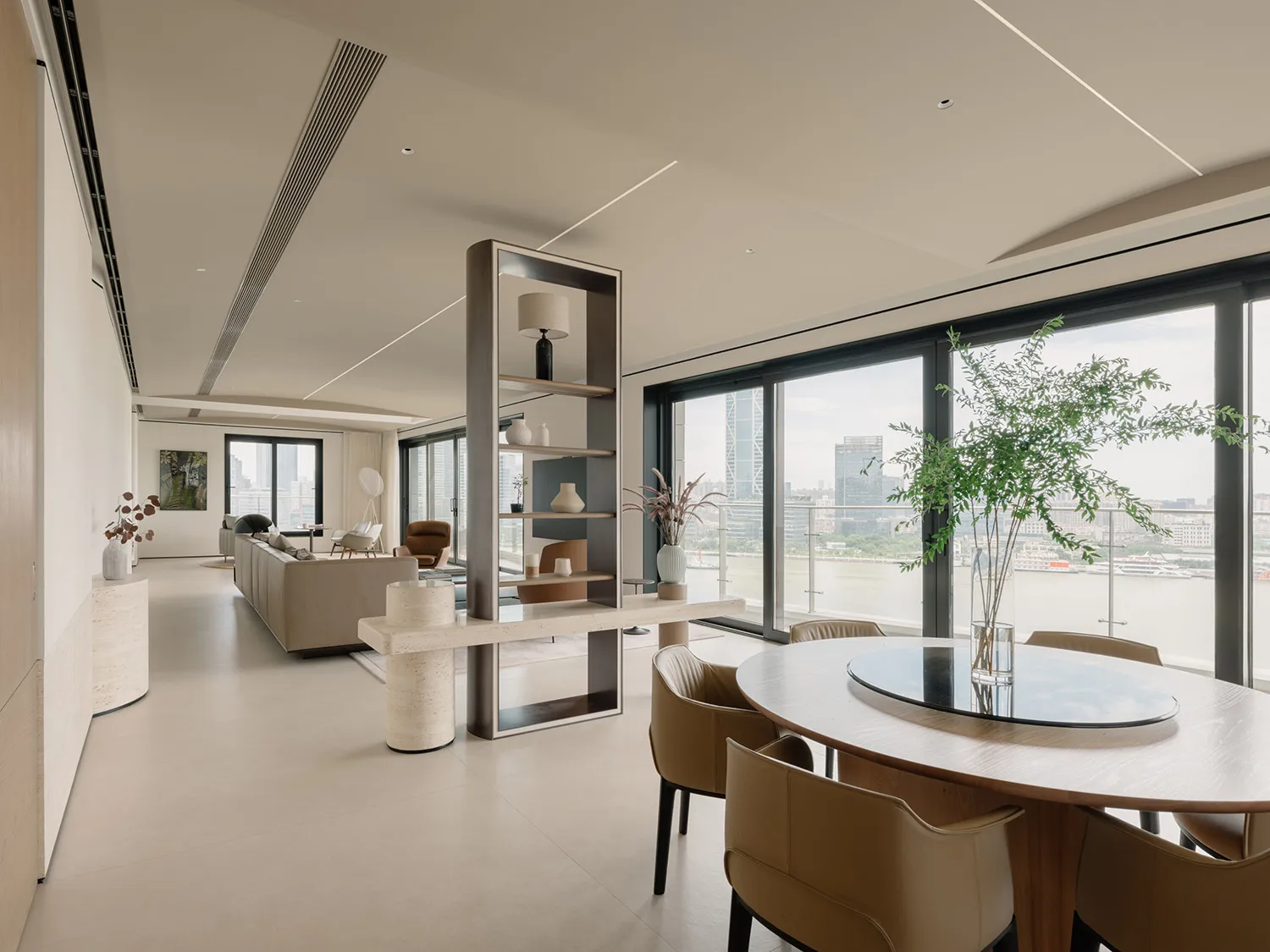 A modern, neutral-toned living space featuring a dining table and chairs, a round table, a long sofa, and tall windows with a
