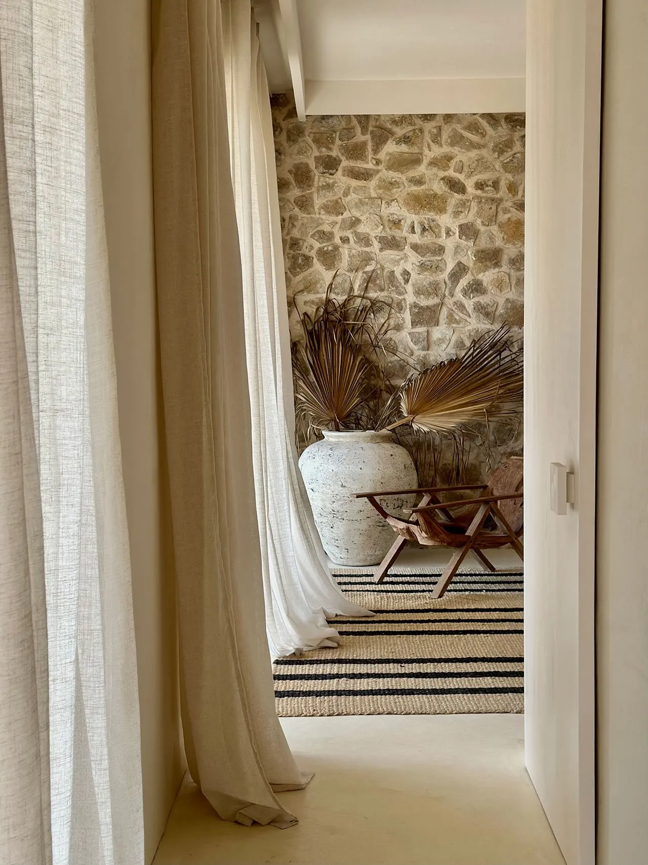 A neutral-toned living room features a stone wall, a large vase with dried palm leaves, a wooden chair, and a striped rug und