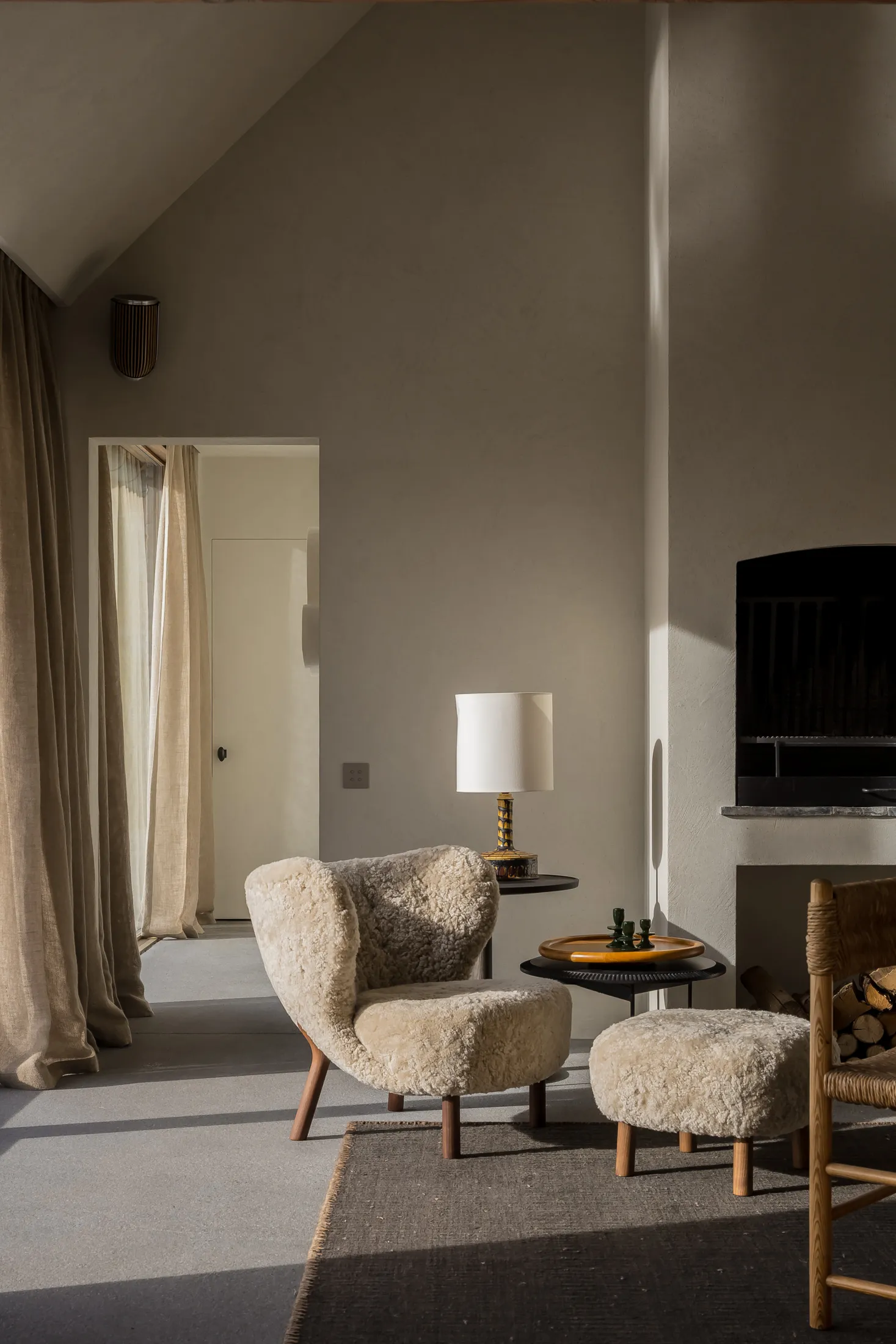 A cozy living room with a sheepskin chair and ottoman, a side table with a lamp, and a fireplace.