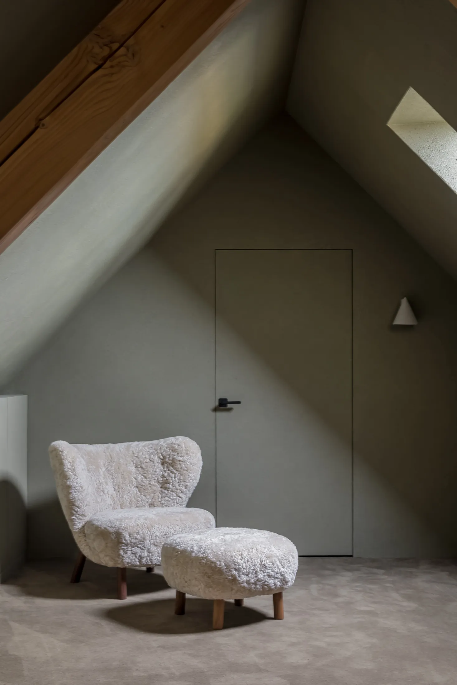 A cozy mid-century living room with a white wool armchair and matching ottoman, set against a muted green wall and slanted ce